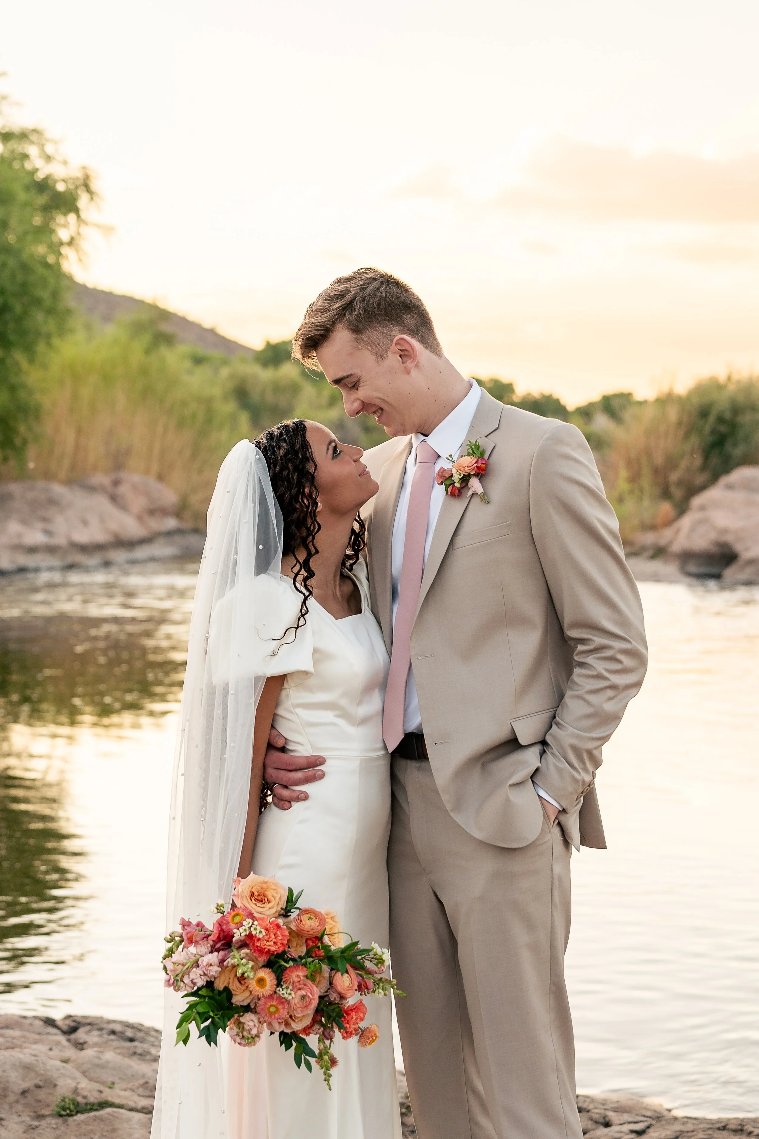 A bride and groom stand close, smiling at each other by a river at sunset, with the bride holding a colorful bouquet of flowers.