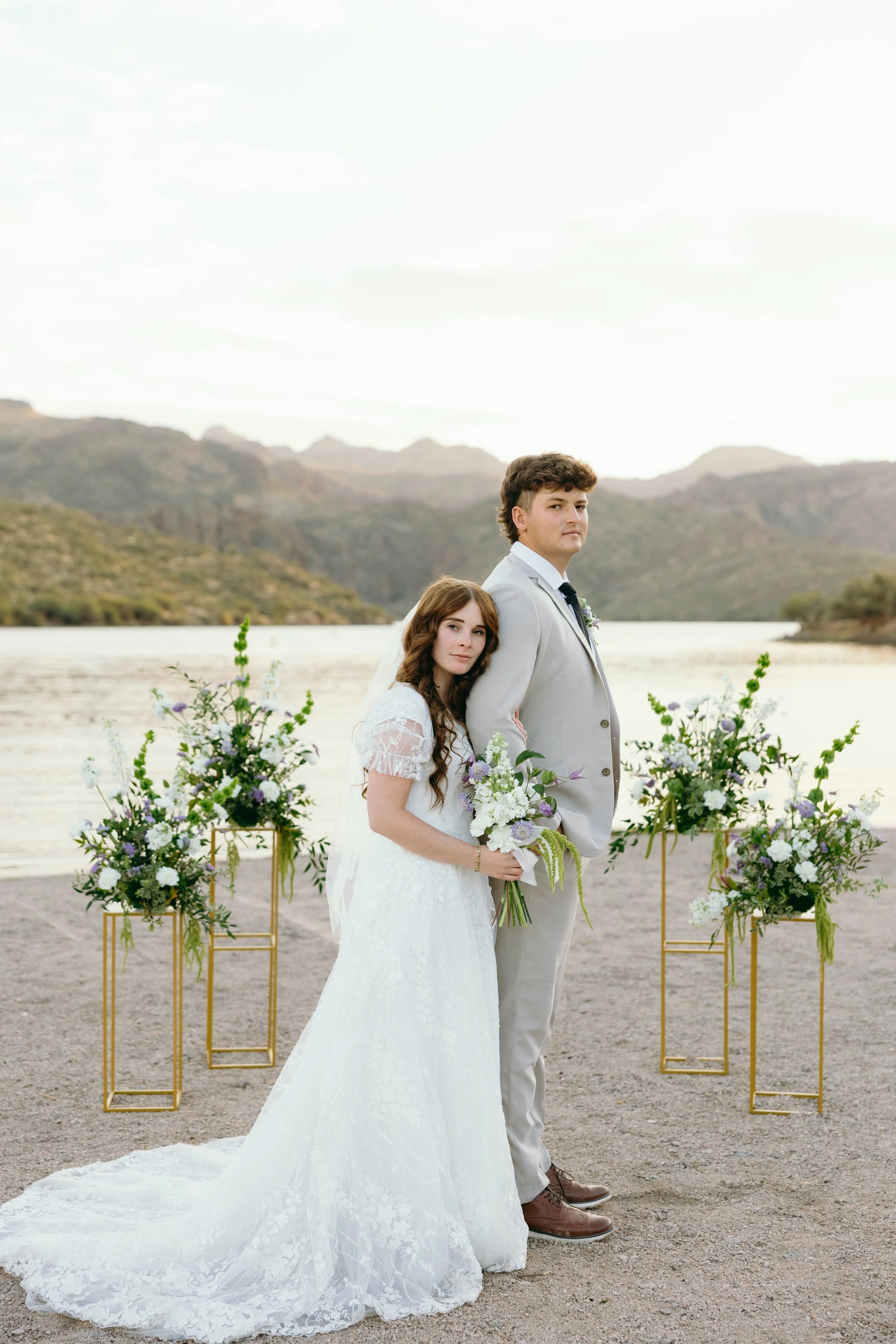 Bride and groom standing together near a river with mountains in the background, decorated with floral arrangements, during a wedding ceremony.