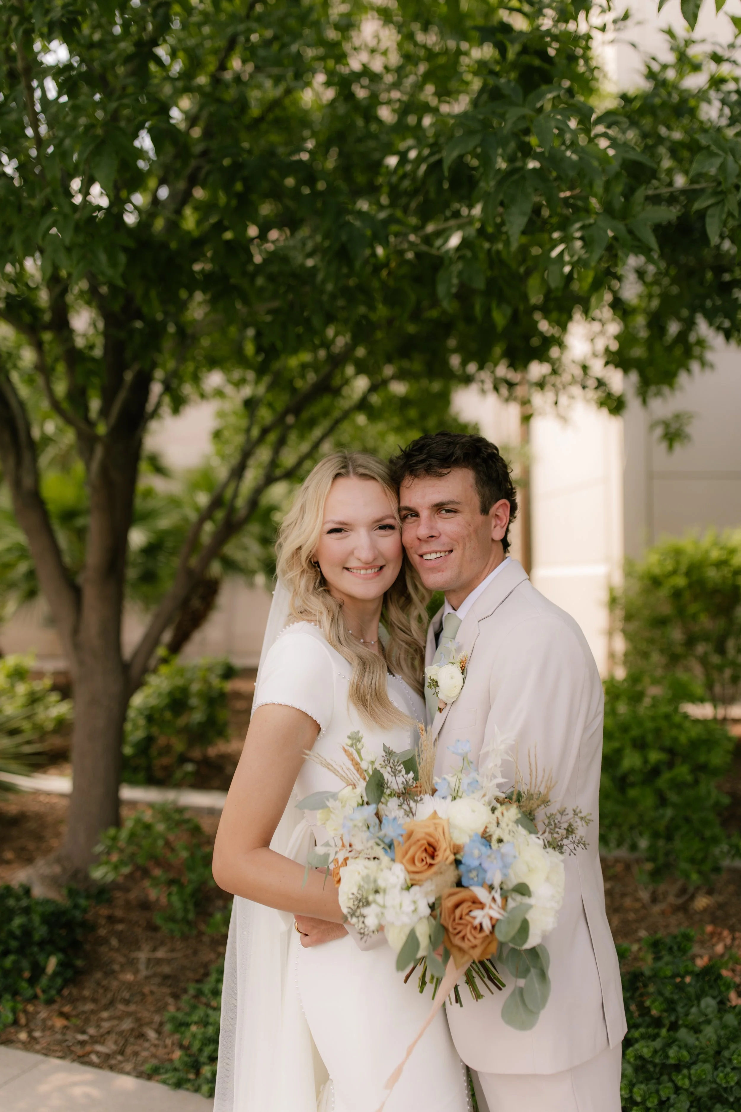A bride and groom smiling and standing close together outdoors. The bride holds a bouquet of roses, greenery, and other flowers. They are dressed in wedding attire, with the bride in a white dress with short sleeves and the groom in a light-colored s
