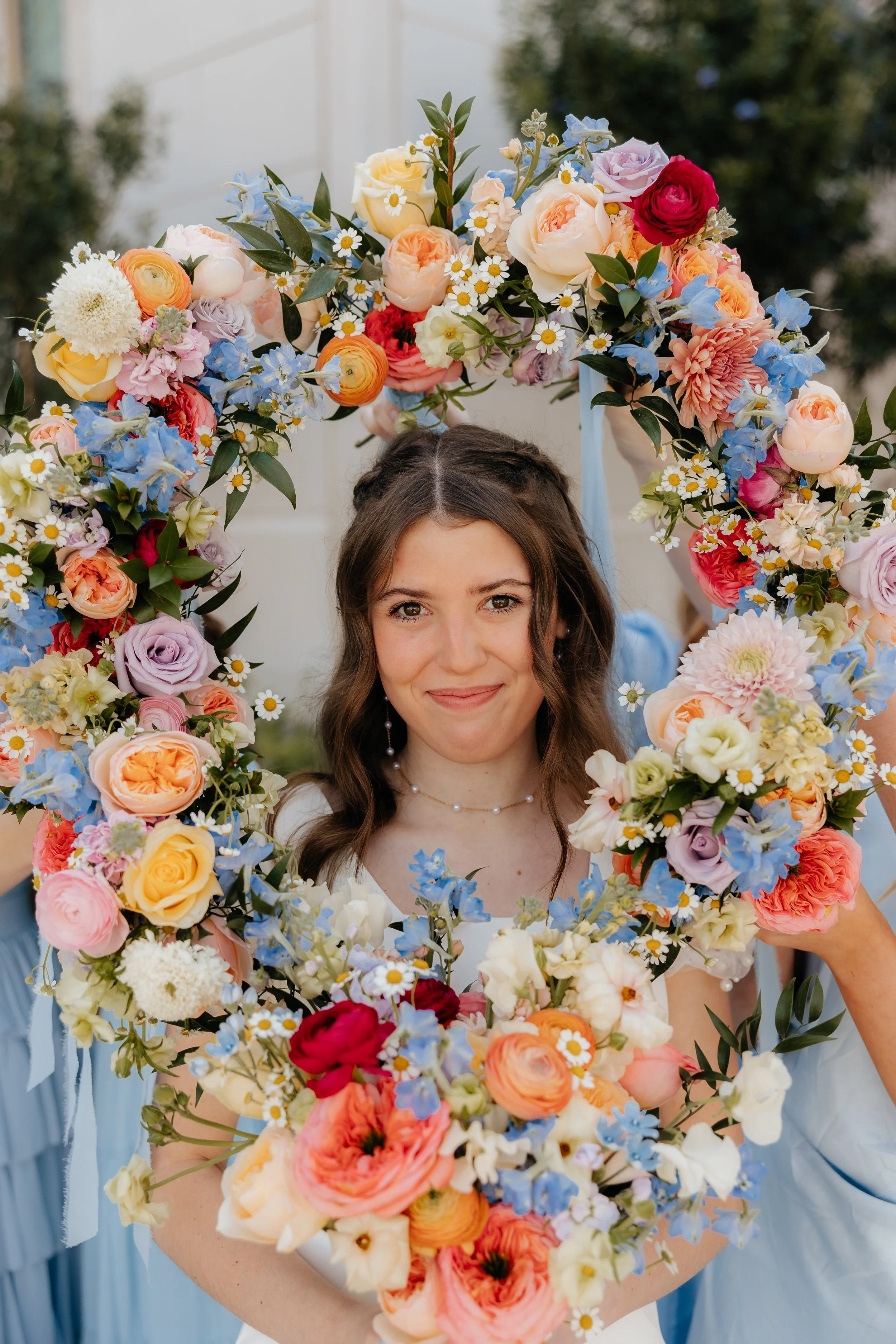 A woman with brown hair and a white dress holding a large floral wreath composed of various colorful flowers including roses, daisies, and delphiniums, smiling at the camera.