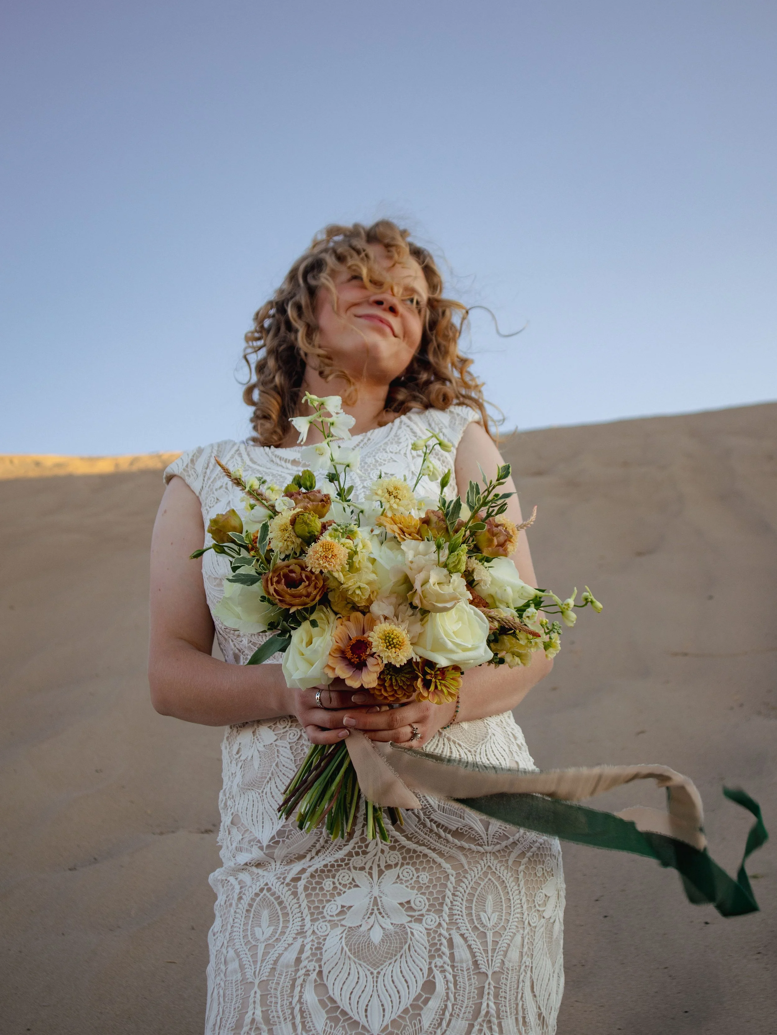 A woman in a white lace dress holding a bouquet of flowers in a desert landscape with a clear blue sky.