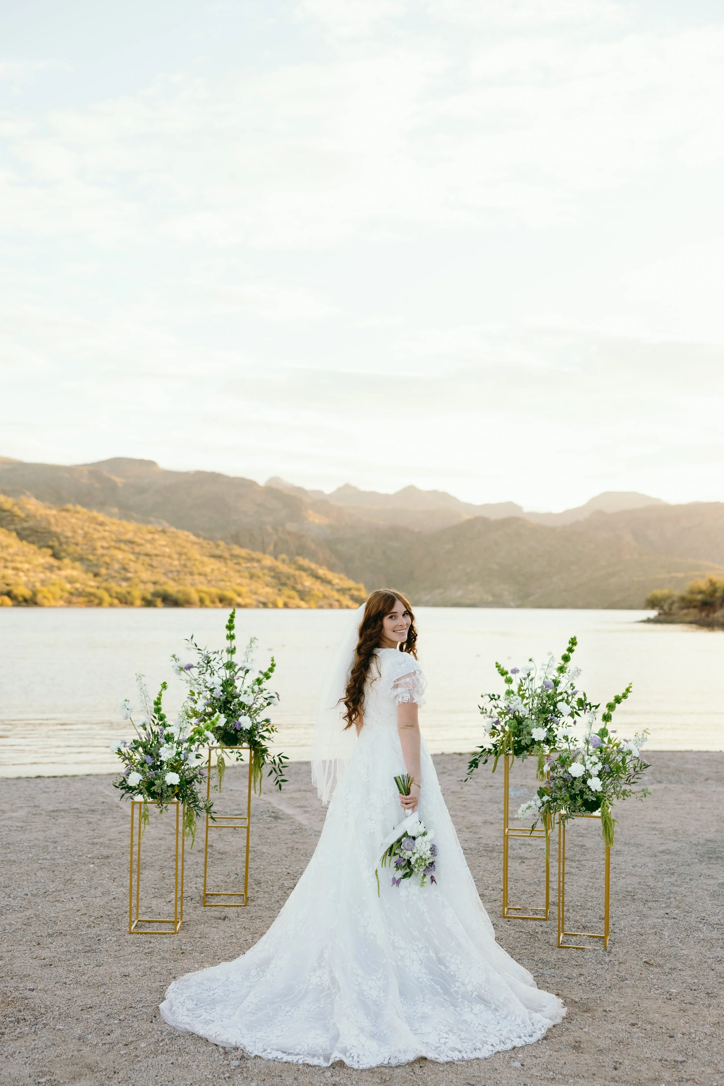 A bride in a white wedding dress holding a bouquet of flowers, standing outdoors near a lake with mountains in the background, surrounded by floral arrangements on golden stands.