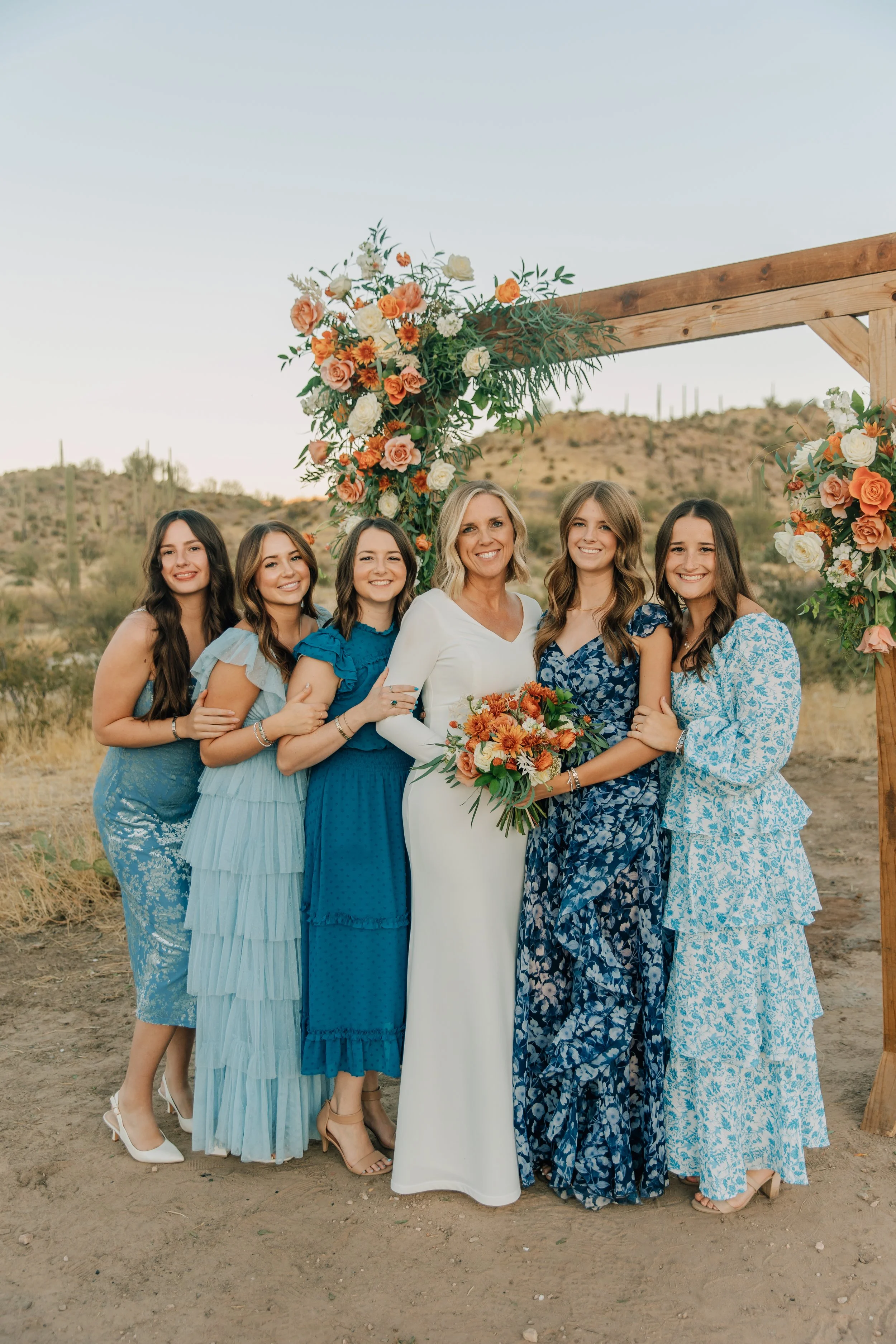 A bride in a white dress holding a bouquet of orange and white flowers, standing with five women in blue dresses under a floral arch at an outdoor desert setting.