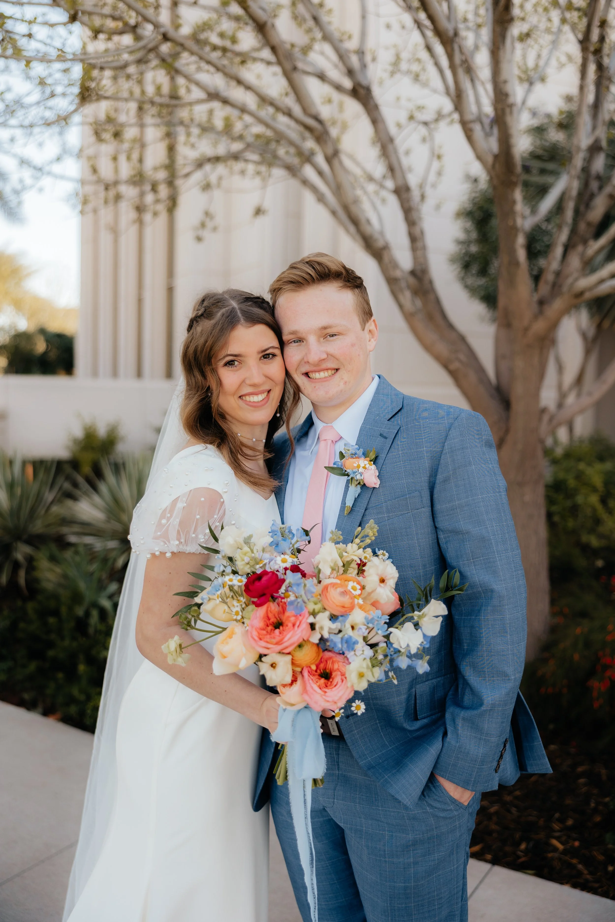 A bride and groom smiling and standing close together outdoors, holding a bouquet of colorful flowers, with a tree and building in the background.