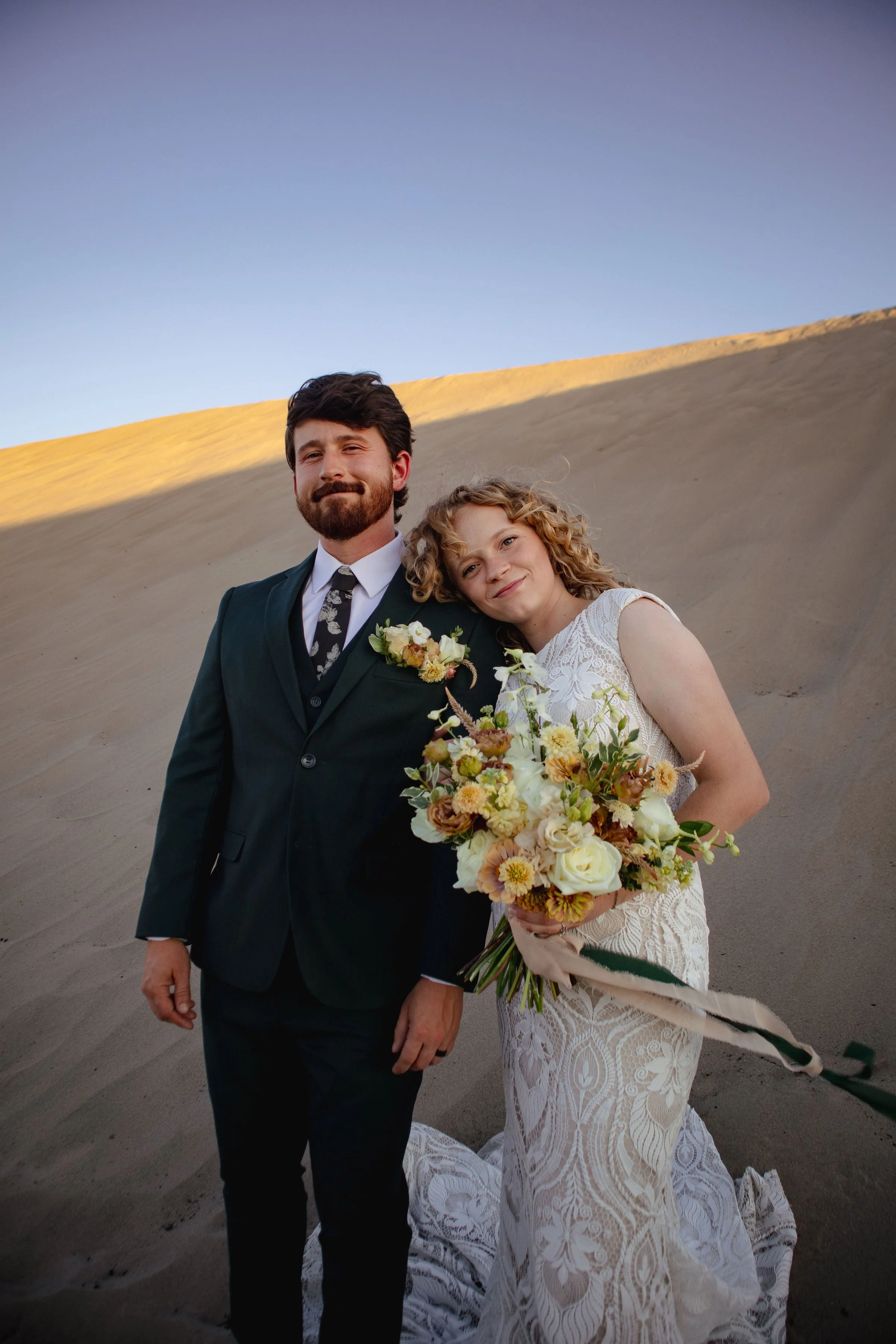 A bride and groom standing together outdoors, with a sandy hill and a clear sky in the background. The bride is holding a bouquet of flowers and leaning her head on the groom's shoulder. The groom is dressed in a dark suit, white shirt, and patterned tie.