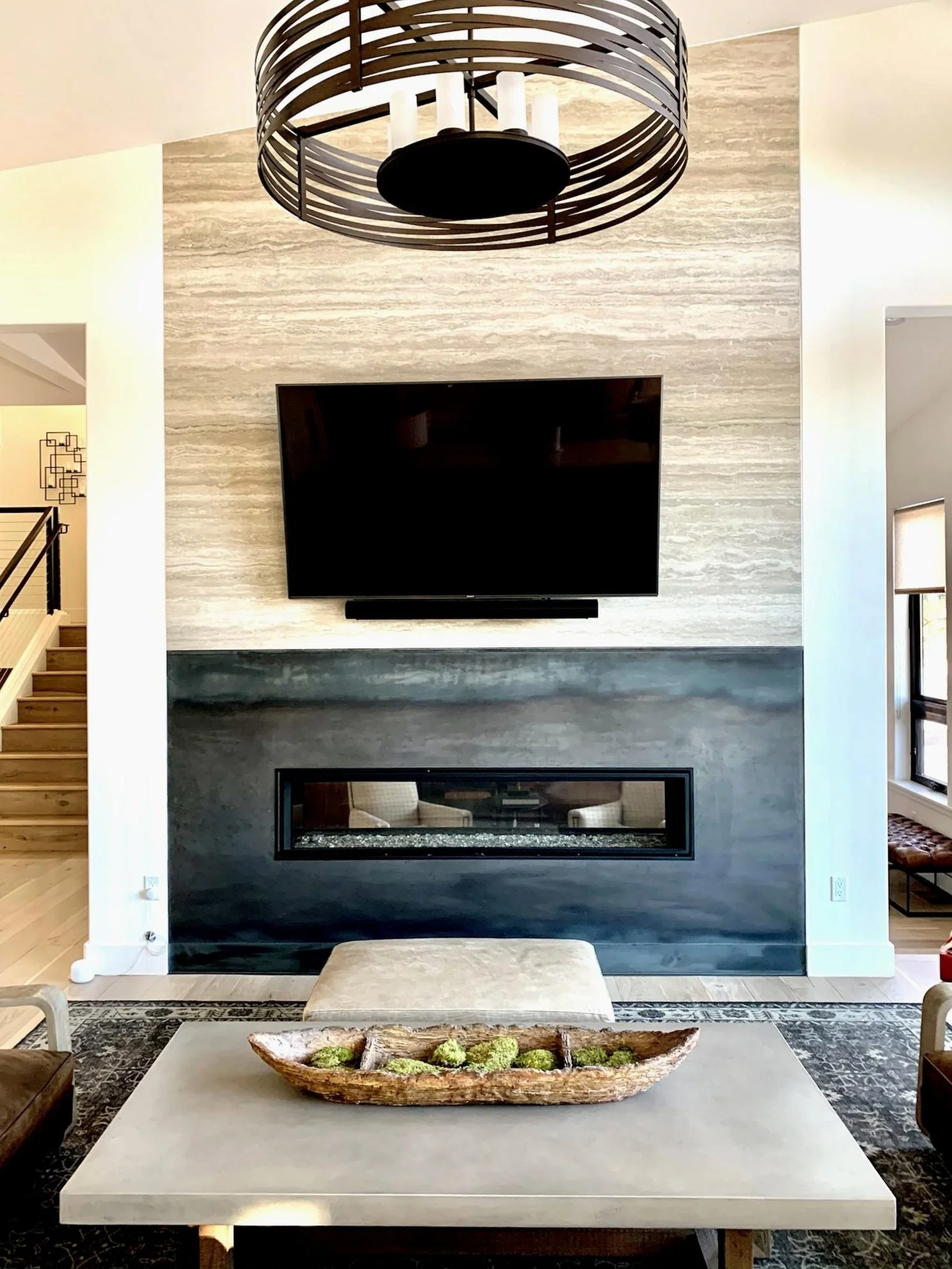 Living room with a wall-mounted TV above a modern fireplace, a rectangular coffee table with green moss and wood decor, and a circular chandelier hanging from the ceiling.