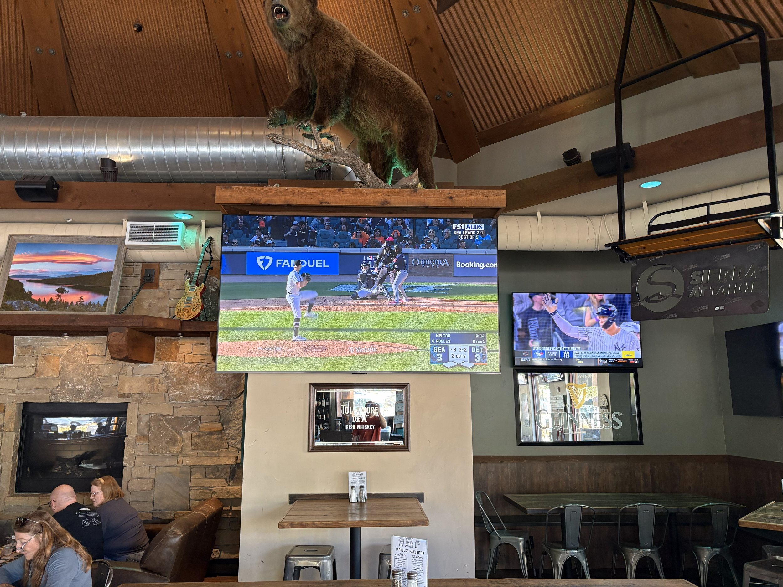 Inside a sports bar with multiple TV screens showing a baseball game, a mounted bear on a wooden shelf above the TVs, a stone fireplace, and people sitting at tables.
