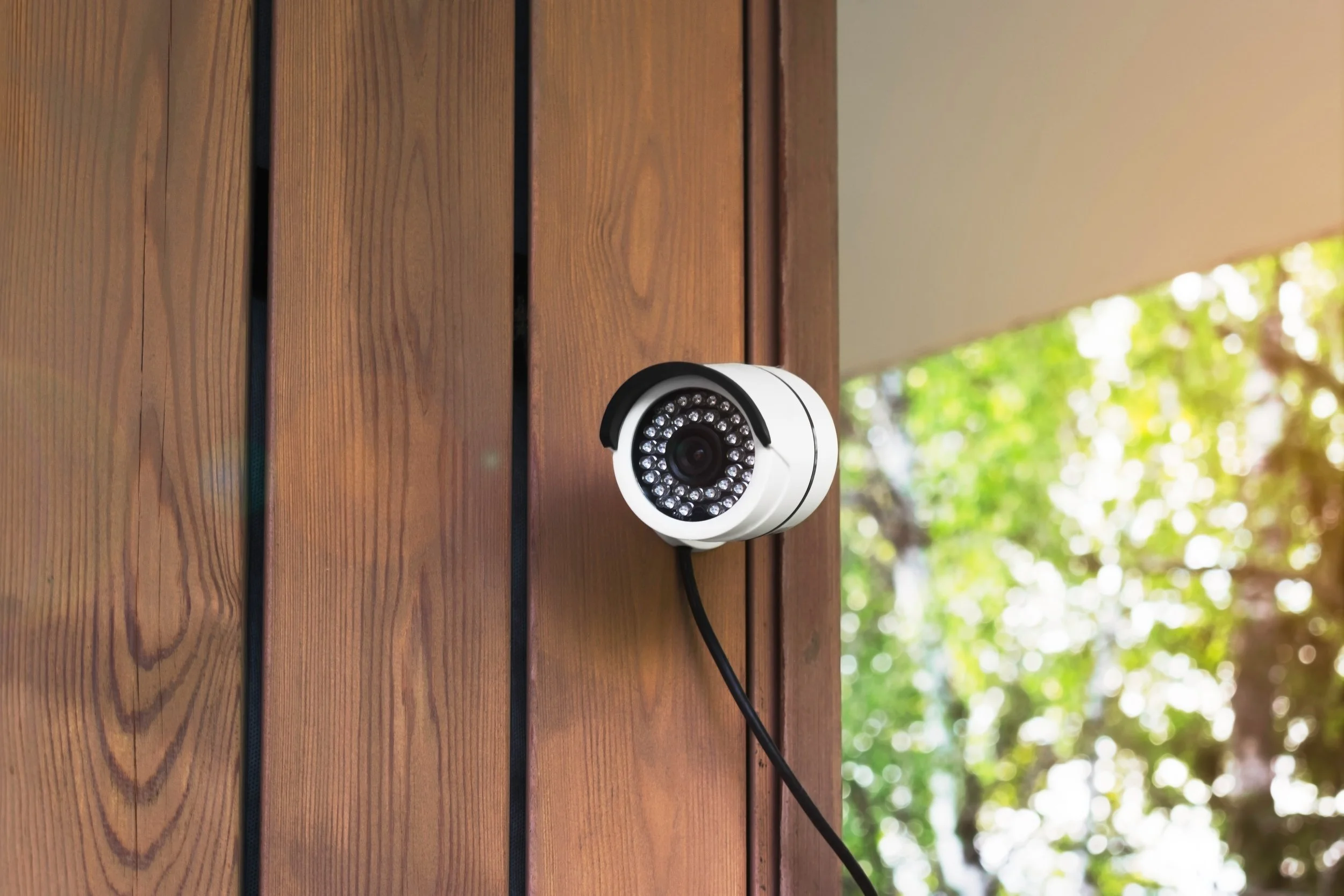 Security camera mounted on wooden wall with trees and sunlight in background.