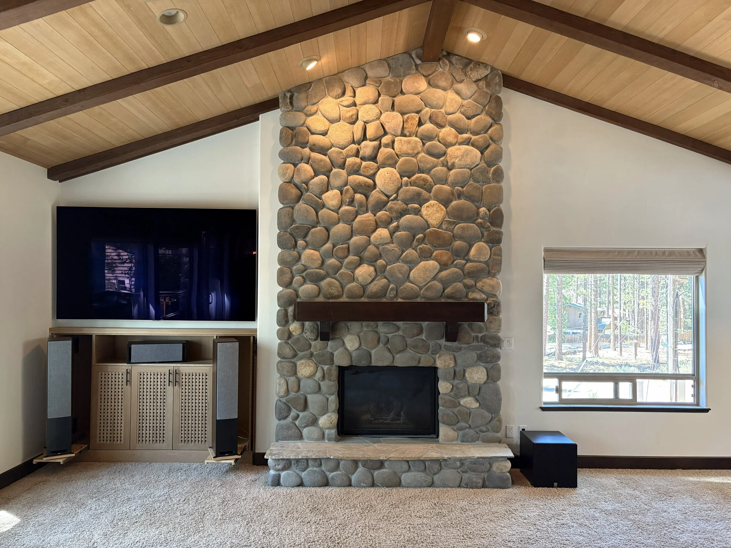 Living room with a stone fireplace, flat-screen TV, built-in audio system, window showing outdoor trees, and sloped wooden ceiling.