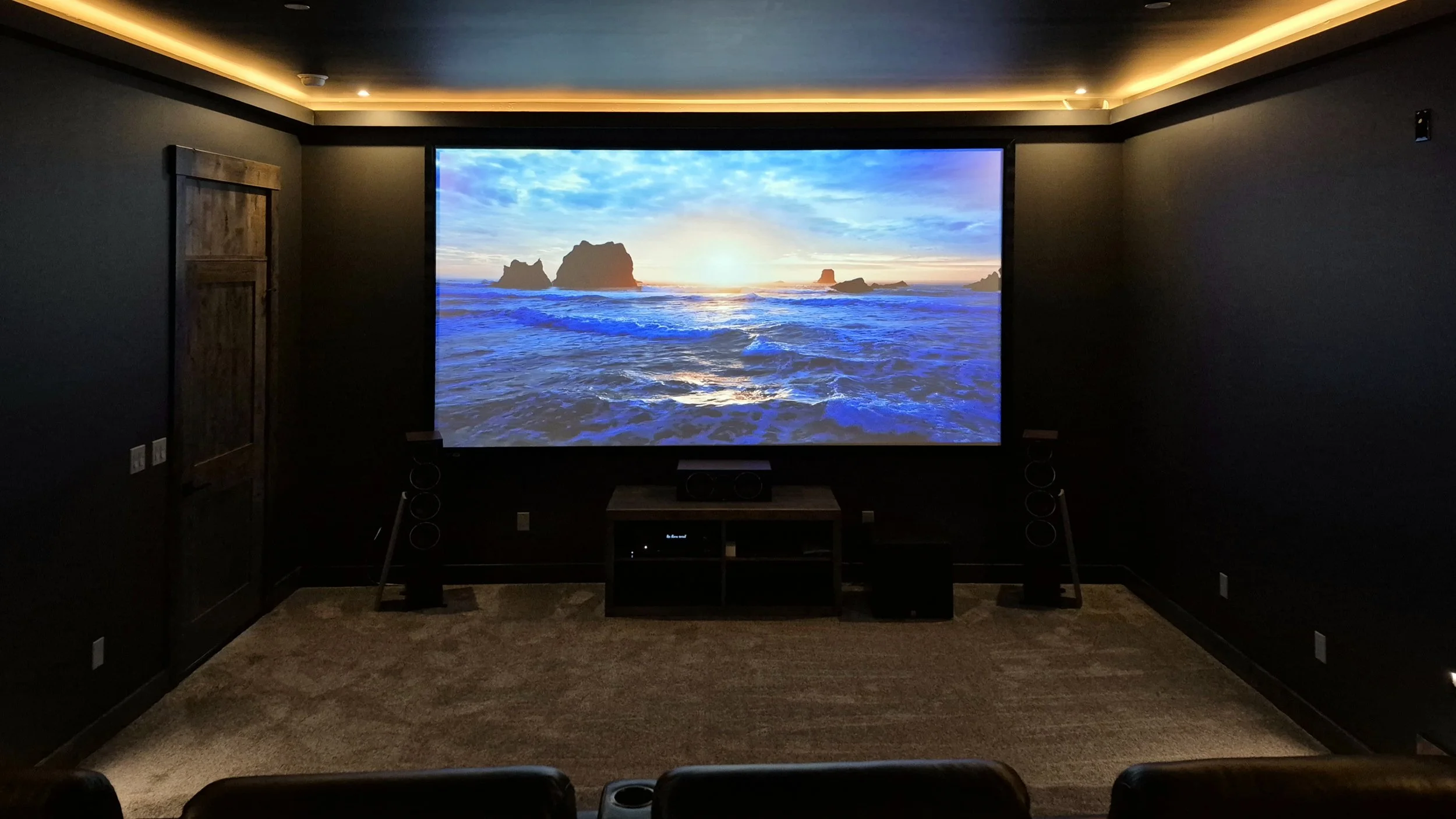 Home theater room with a large screen showing a sunset over ocean waves and rock formations.