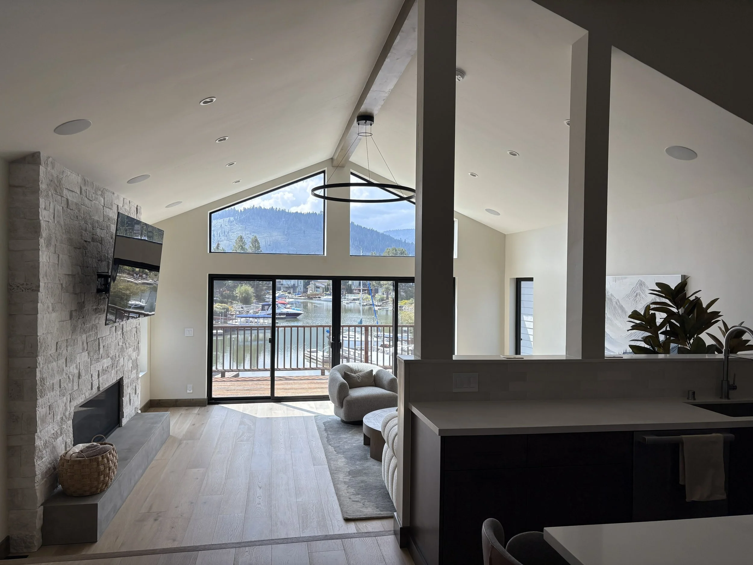 Living room with large windows overlooking a water harbor and mountains, stone fireplace, wall-mounted TV, cream-colored armchair, sliding glass door leading to outdoor deck, modern lighting, and partial view of kitchen with countertop and sink.