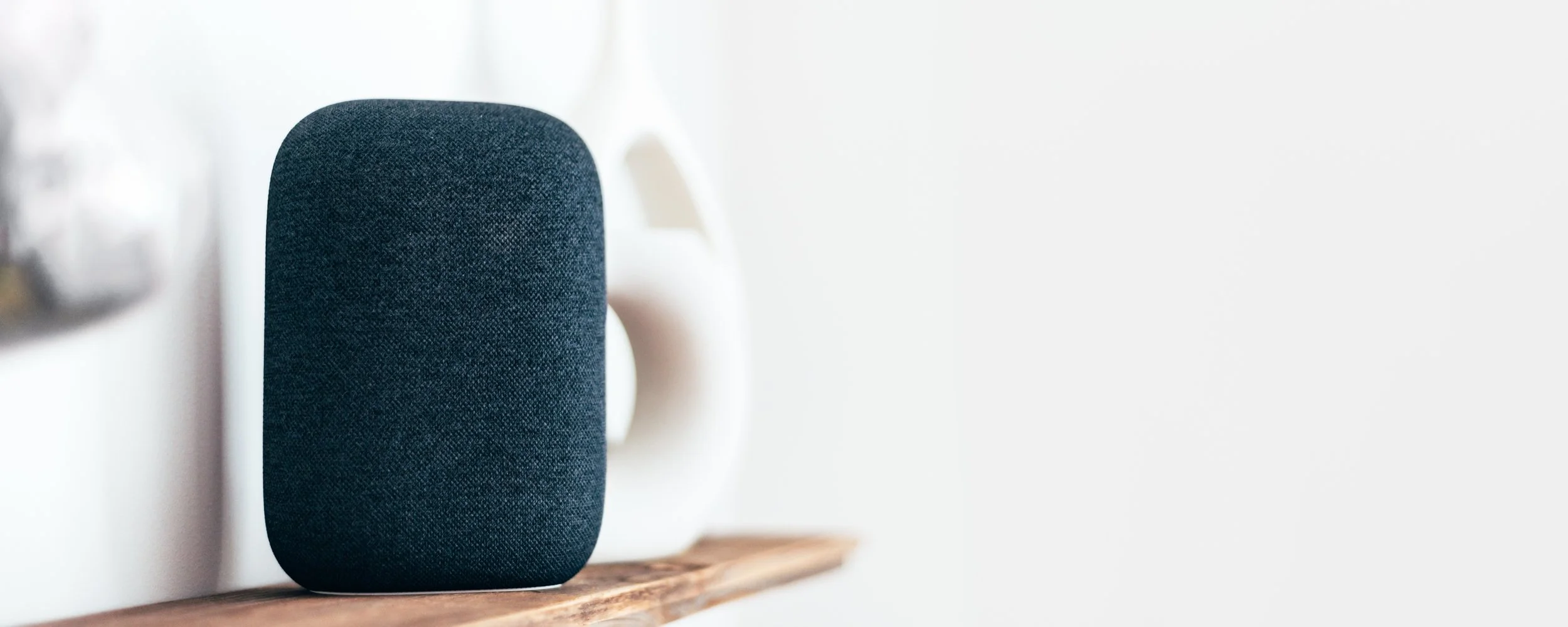 Smart speaker with a fabric cover sitting on a wooden shelf next to a white device against a white wall.