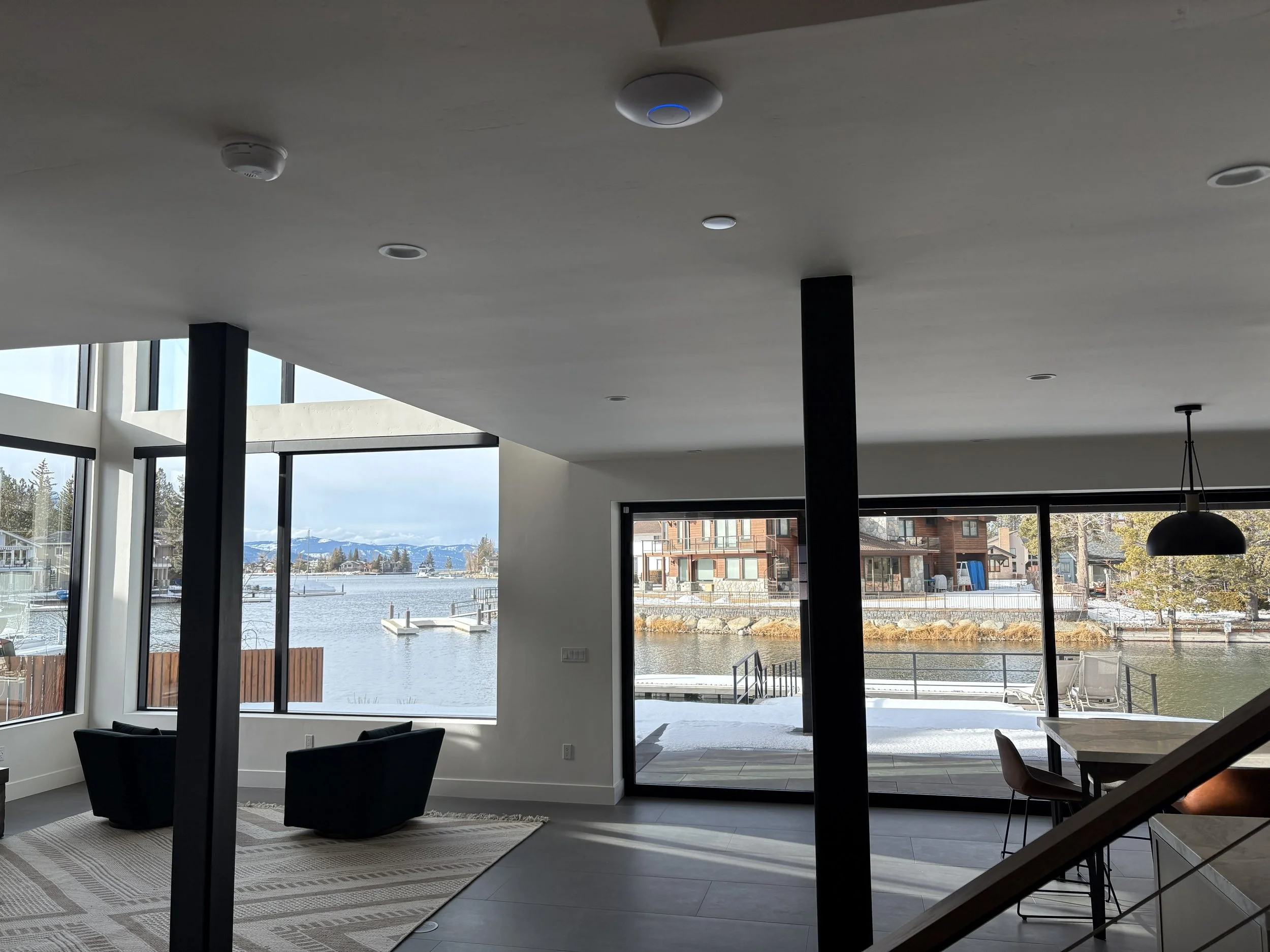 Modern living room with large windows overlooking a waterfront. The room has black armchairs, a patterned rug, and a kitchen counter with chairs. Outside, there is snow on the ground, a deck with outdoor furniture, and houses across the water with snow-capped mountains in the distance.