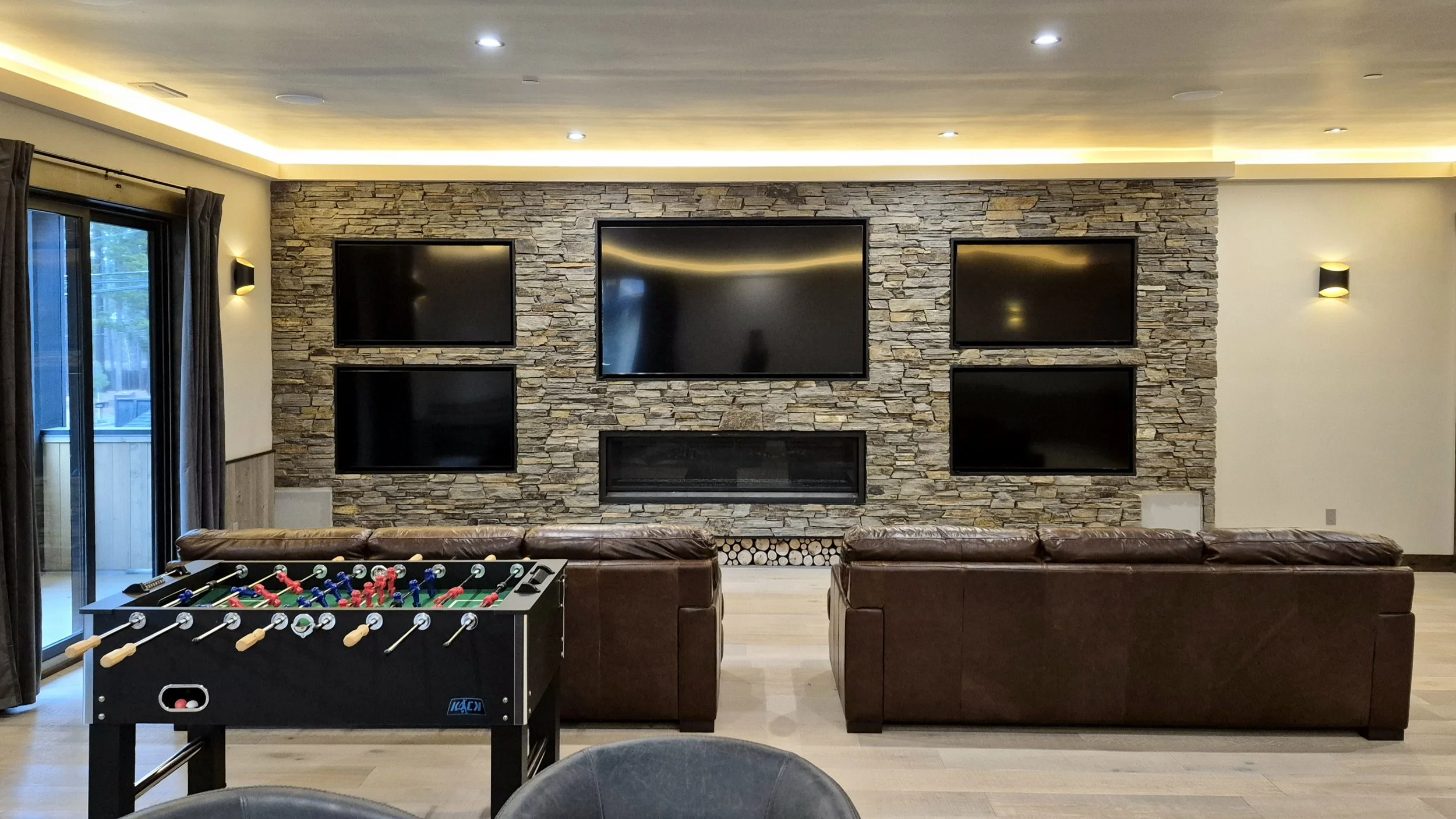 Living room with stone accent wall, four flat-screen TVs, leather sofas, and a foosball table.
