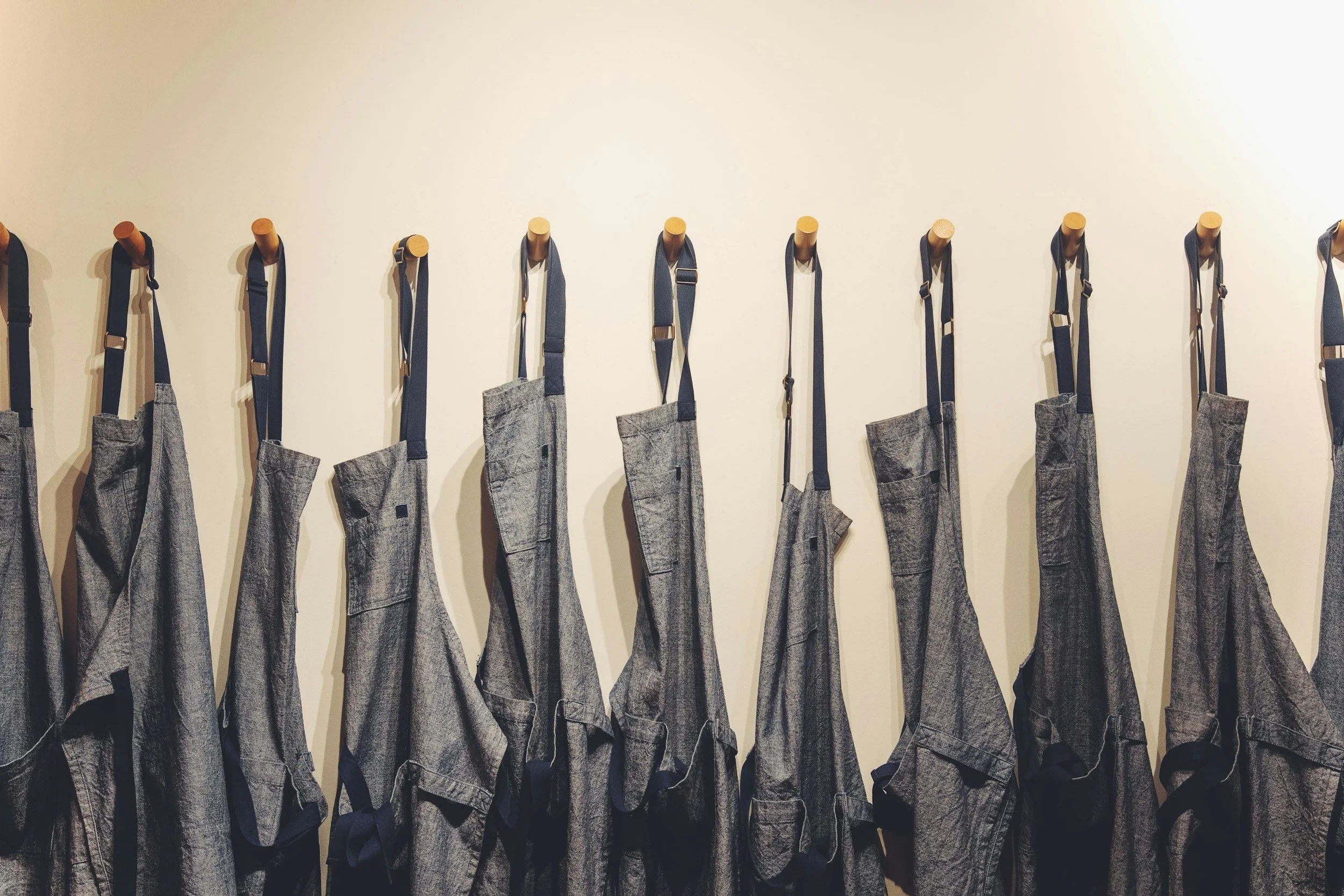 Multiple gray aprons hanging on wooden pegs on a white wall.