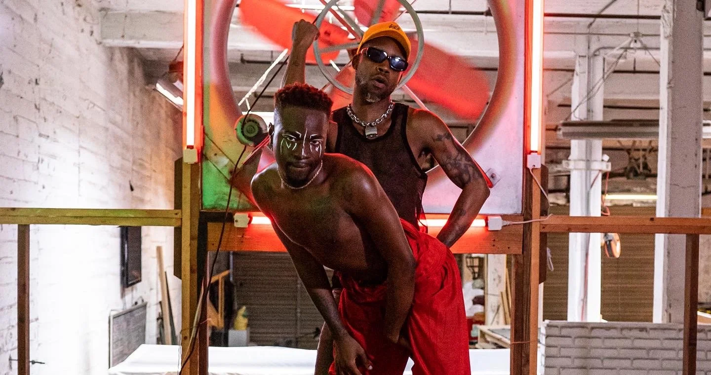 Two shirtless men with tattoos and bold makeup in an industrial setting with wooden framework and neon lights. One man is leaning forward with red pants, and the other stands behind with sunglasses, a black tank top, and chains.