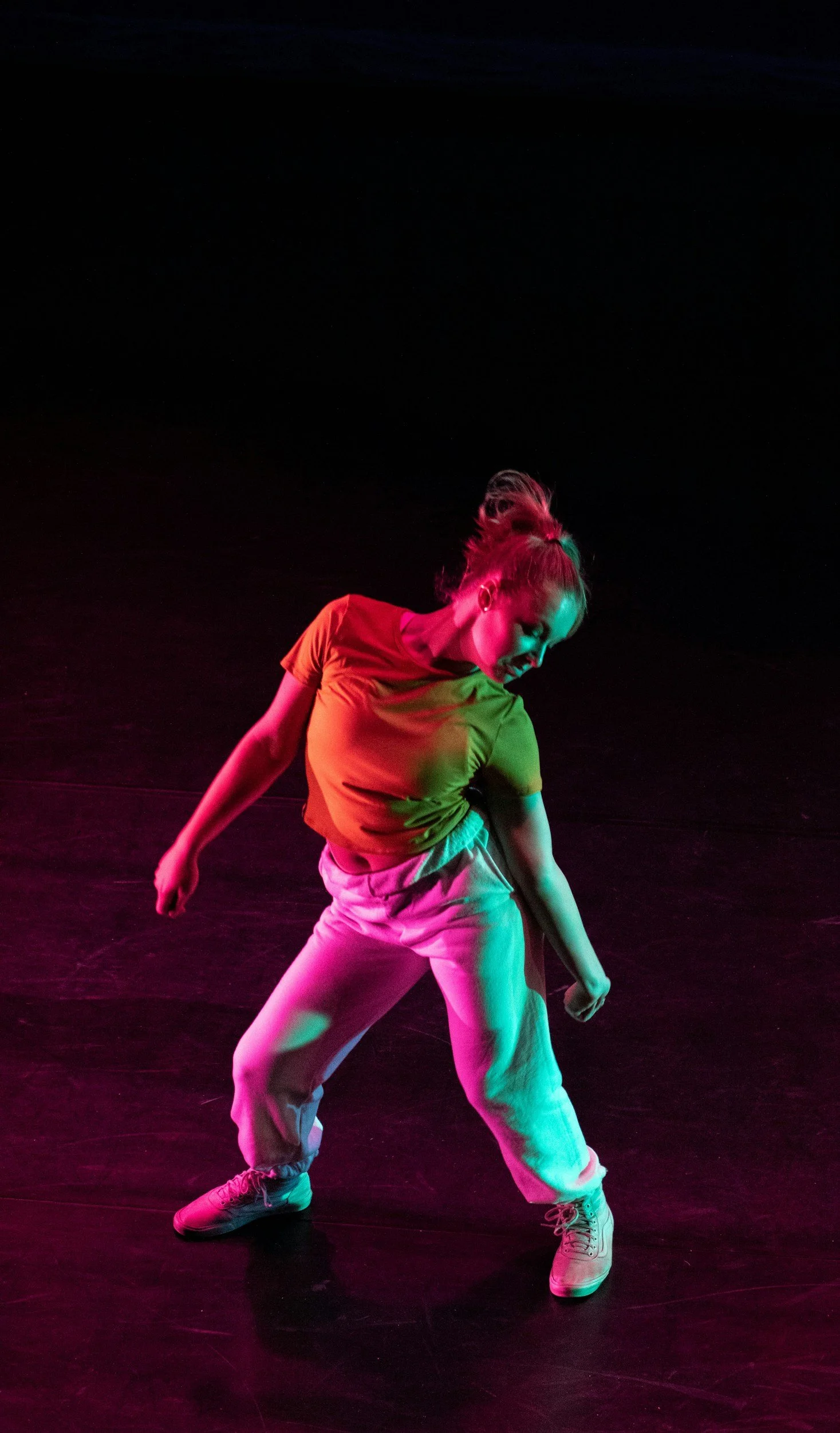 A young woman dancing on a dark stage, illuminated by colorful pink and green lighting, wearing a yellow top, white pants, and sneakers.