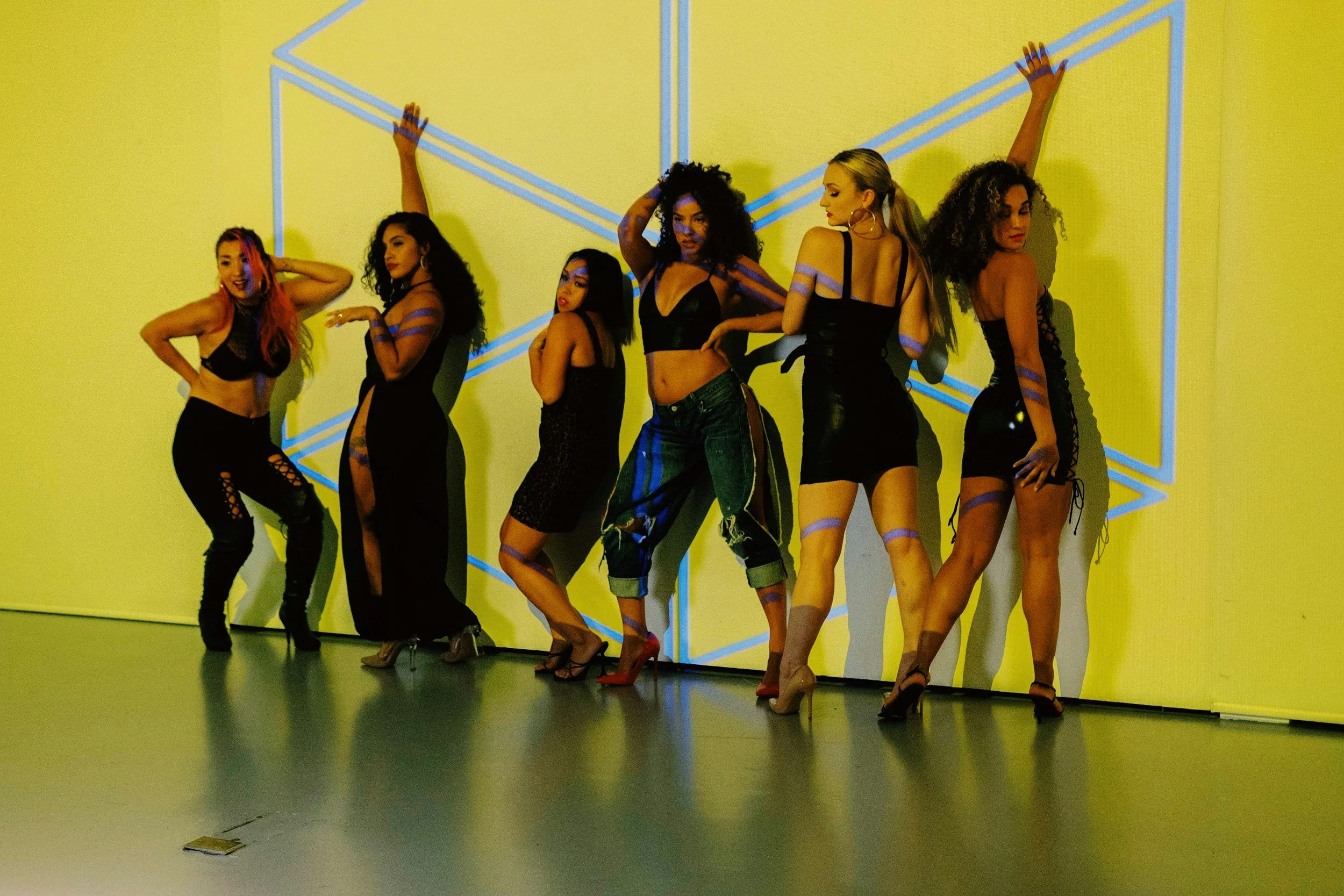 Six women posing in front of a yellow wall with blue geometric lines, wearing black outfits and high heels.