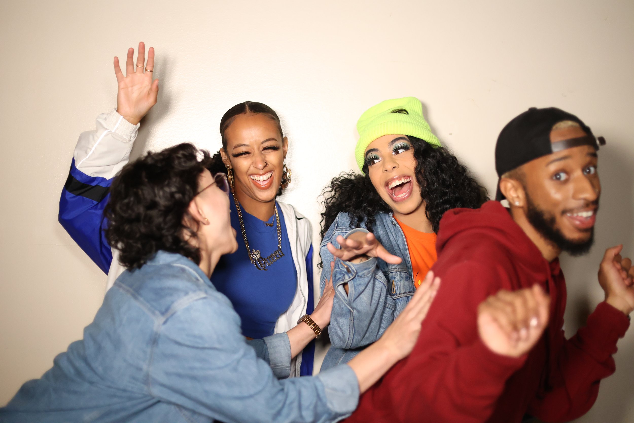 Four young adults laughing and joking together, posing playfully against a plain wall.