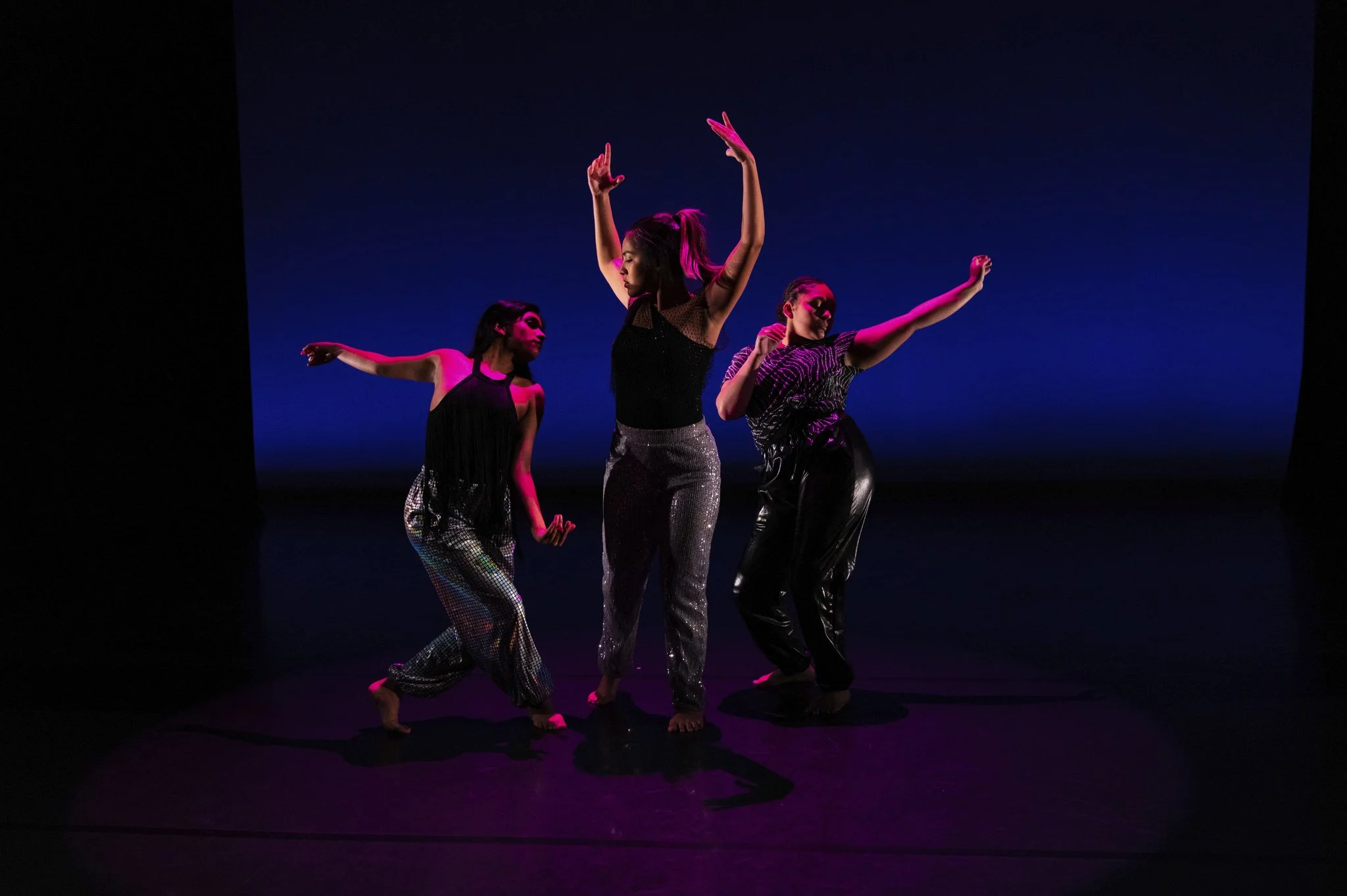Three women in shiny, colorful dance outfits performing on stage with blue background and purple lighting.