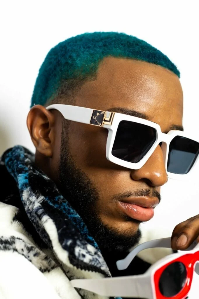 Close-up of a man with vibrant turquoise hair, dark sunglasses, a well-groomed beard, and wearing a patterned hoodie with black, white, and gray colors.