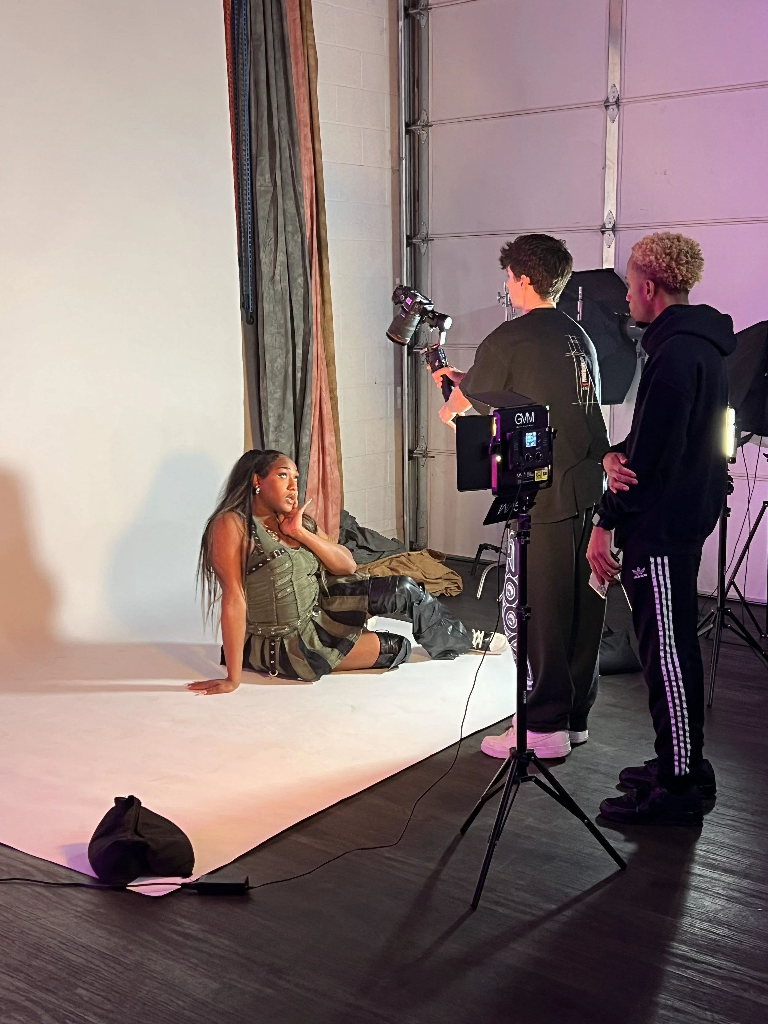 A photoshoot session with a woman sitting on a white backdrop in a studio, being photographed by two men, one holding a camera and the other observing, with studio equipment and a garage door in the background.