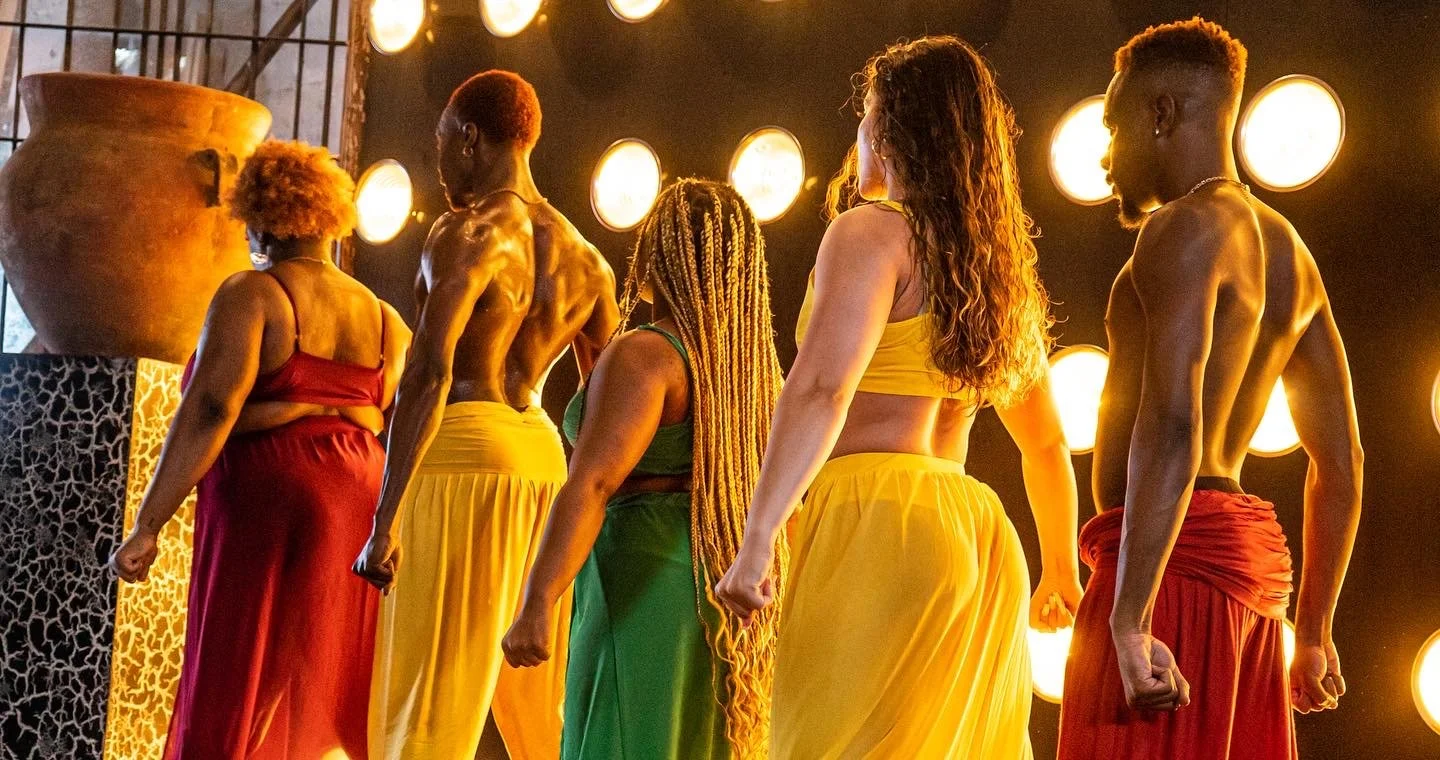 A group of five diverse performers with muscular builds, dressed in colorful costumes, standing on stage in a line with dramatic lighting and large round lights in the background.