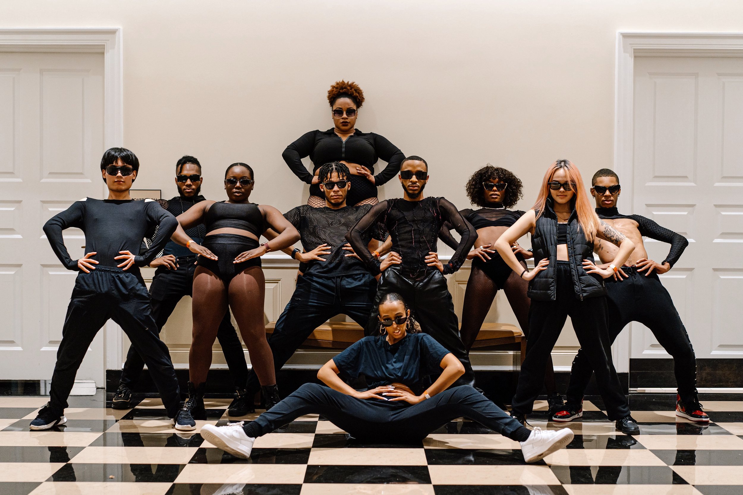 Group of nine diverse individuals in black athletic and stylish clothing, posing confidently in a room with checkered black and white floor and white paneled walls, striking various powerful poses.