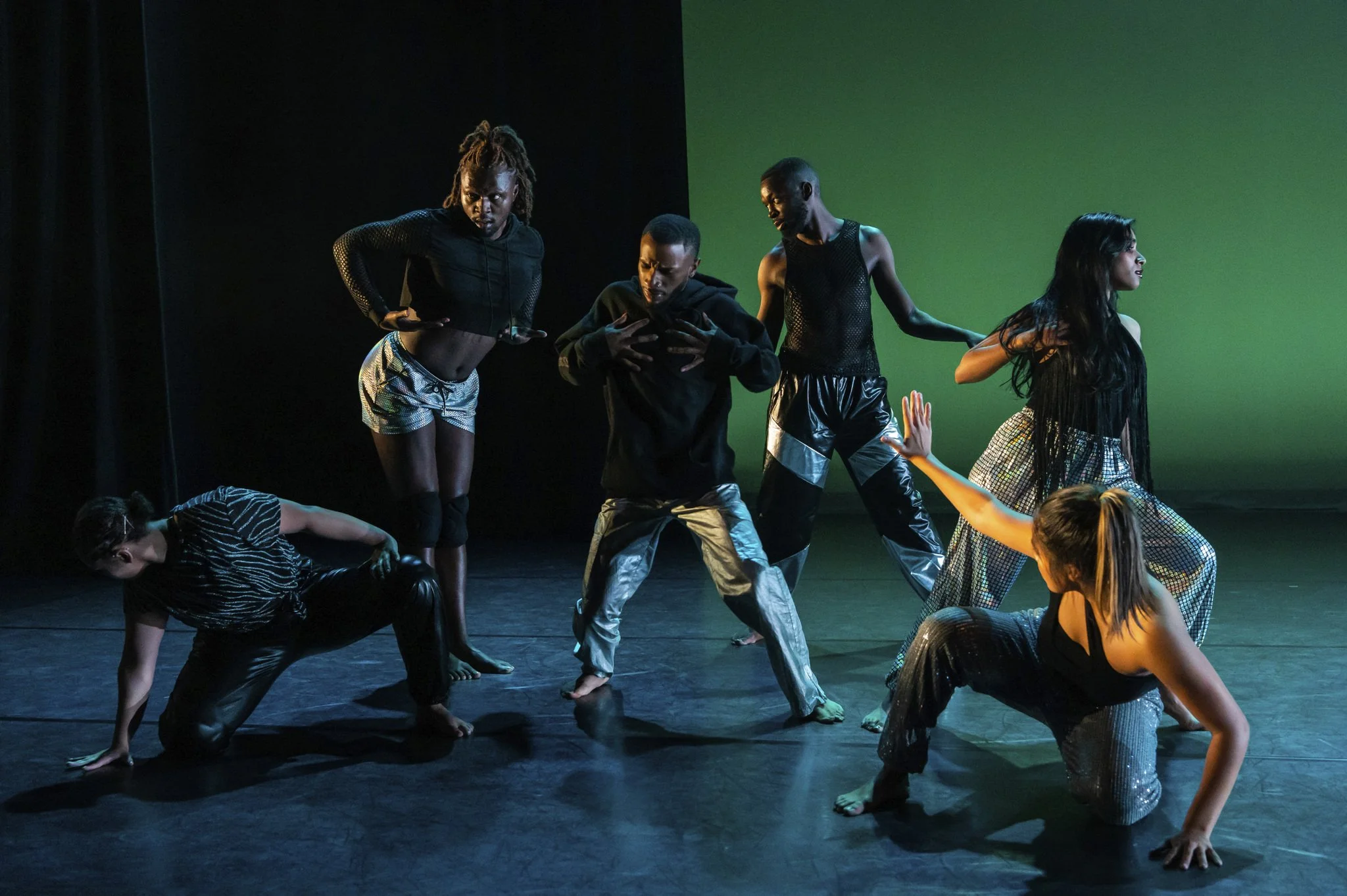 Group of six diverse dancers performing on stage with black and green backgrounds, dressed in black and metallic dance attire.