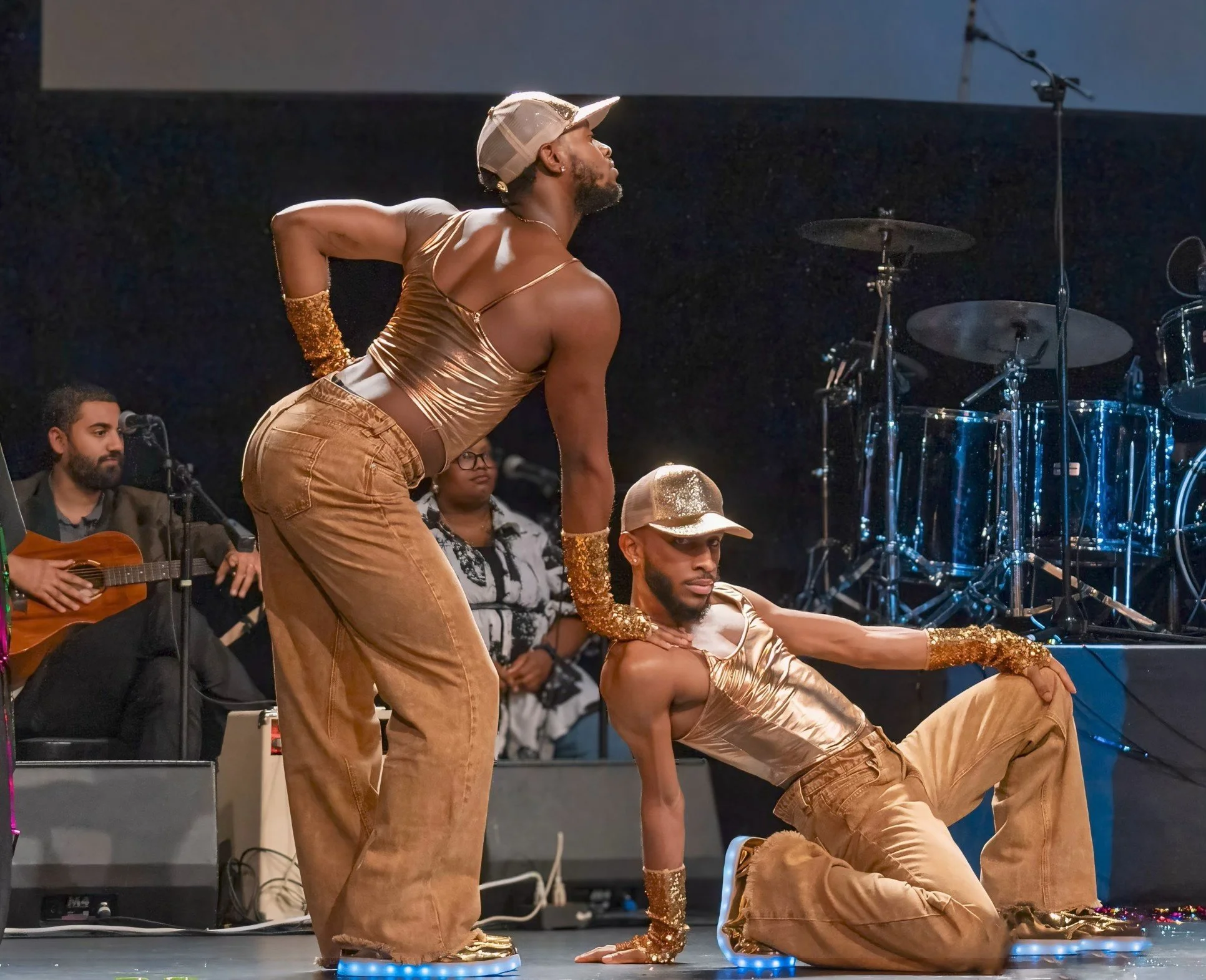 Two dancers in gold tops and jeans perform on stage with musicians in the background, including a person playing guitar and a woman standing near drums.
