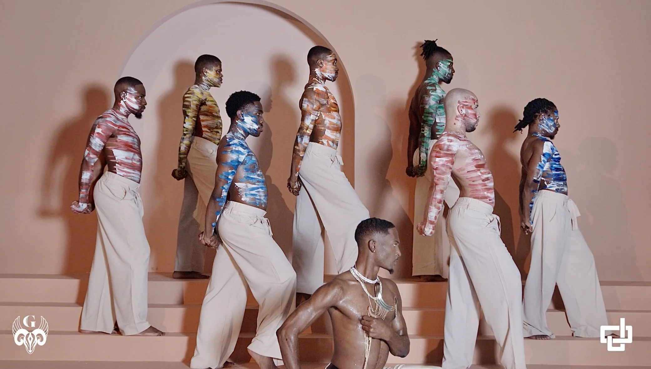 A group of people with painted bodies standing on steps in front of a beige wall. One person is shirtless, wearing necklaces, sitting on the steps, while the others are standing, dressed in loose beige pants.