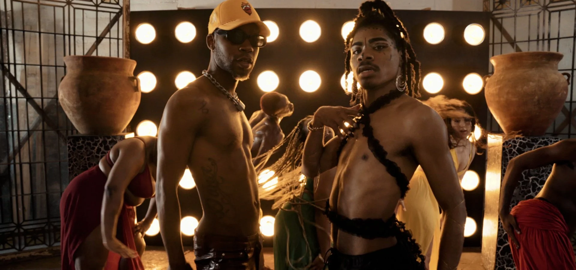Two shirtless men with tattoos and dreadlocks stand in front of a black backdrop with illuminated circular lights. The man on the left wears a beige cap and sunglasses, while the man on the right wears dark makeup and large earrings. There are women in colorful skirts around them, some bending and some standing, in a setting with large clay pots and decorative panels.