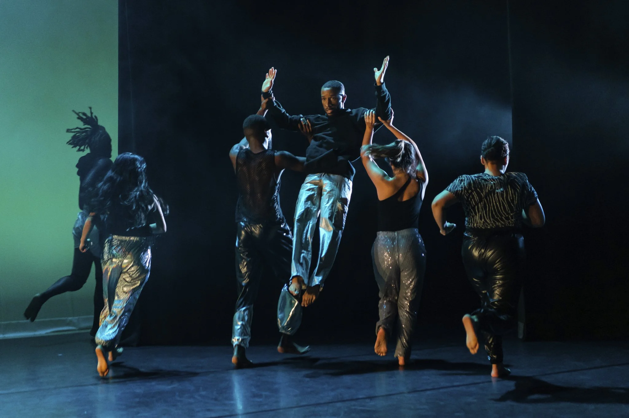 Dance performance on stage with six dancers, one male center, some with arms raised, dressed in shiny pants and black shirts, with dramatic lighting and a dark background.