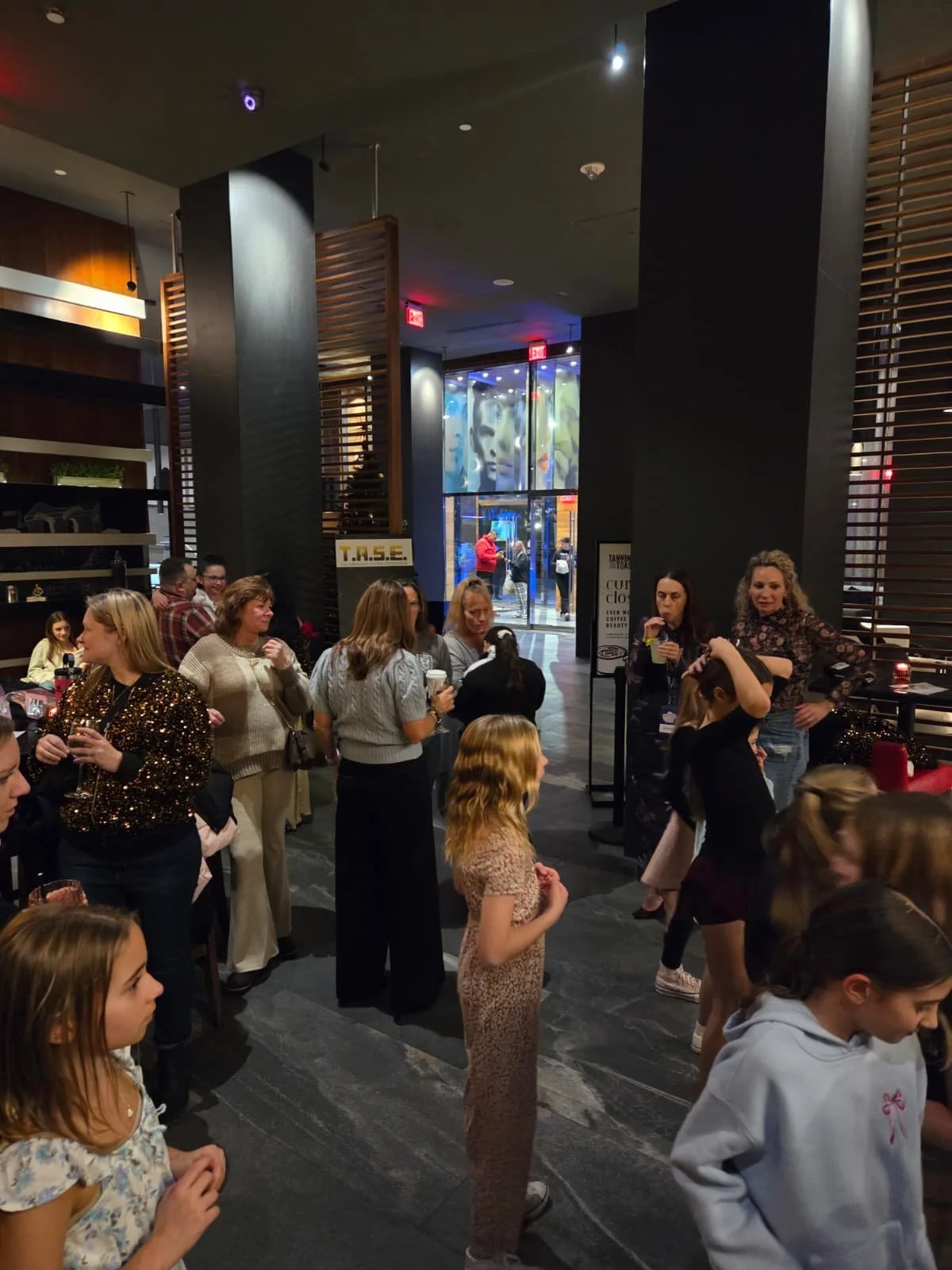 People gathered inside a modern restaurant or bar, socializing, standing near the entrance, with some holding drinks. The decor features dark walls, wooden accents, and a large artistic mural outside visible through glass doors.