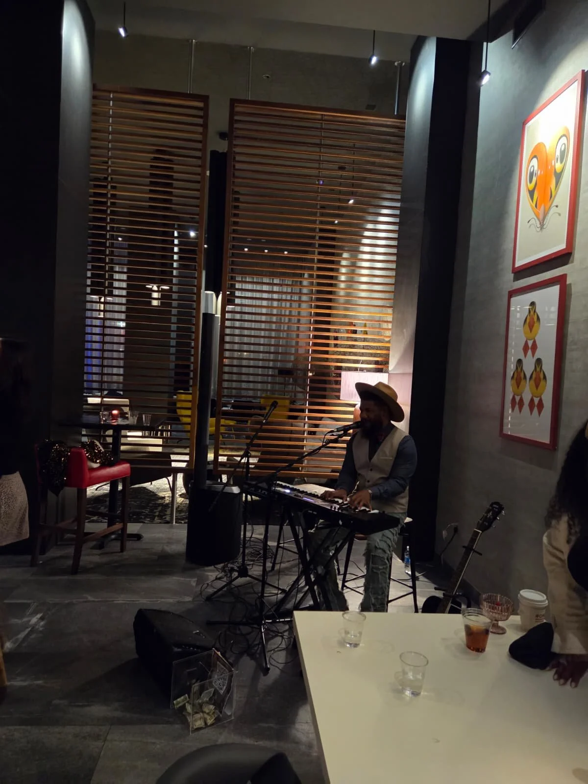 A musician wearing a hat and vest plays a keyboard and sings into a microphone inside a dimly lit restaurant or bar with modern decor, framed art on the wall, and a small table with drinks and a glass of water in the foreground.