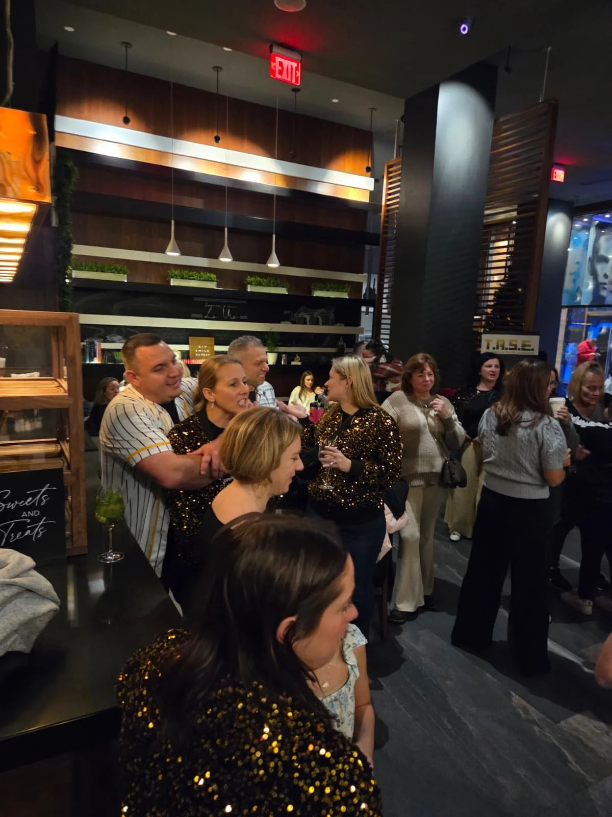 A group of people gathered indoors at a social event, some wearing sparkling outfits, with drinks in hand, in a modern, warmly lit space.