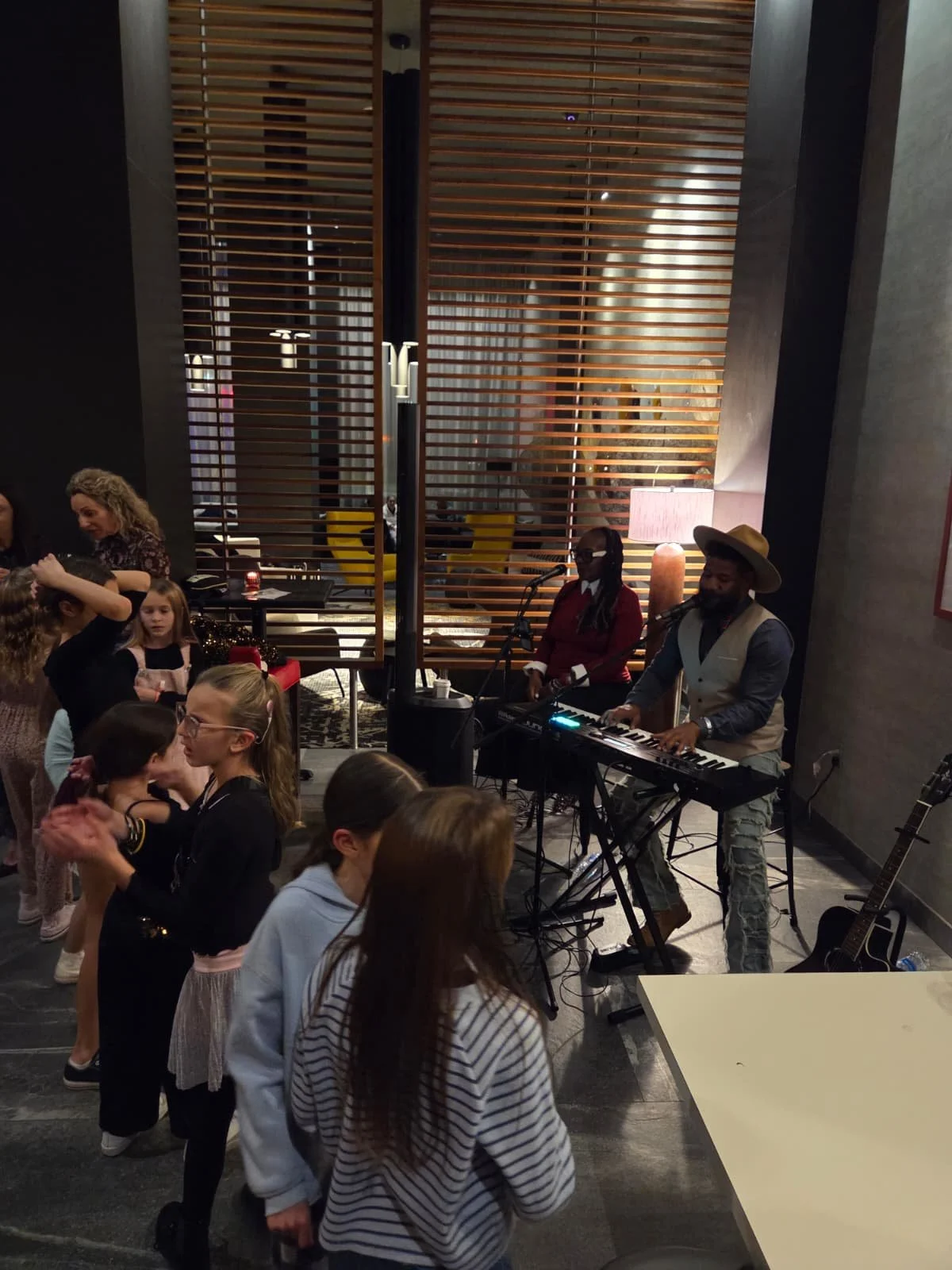 Kids dancing to live music performed by a band with a keyboardist and a singer, in a modern indoor setting.