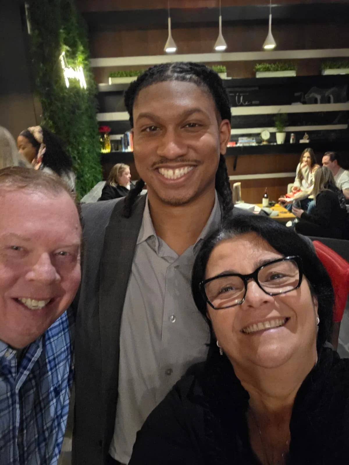 Three people smiling for a photo at a restaurant or cafe, with a man in the middle, a woman with glasses on the right, and a man on the left. The background shows other guests seated and decorative shelves.