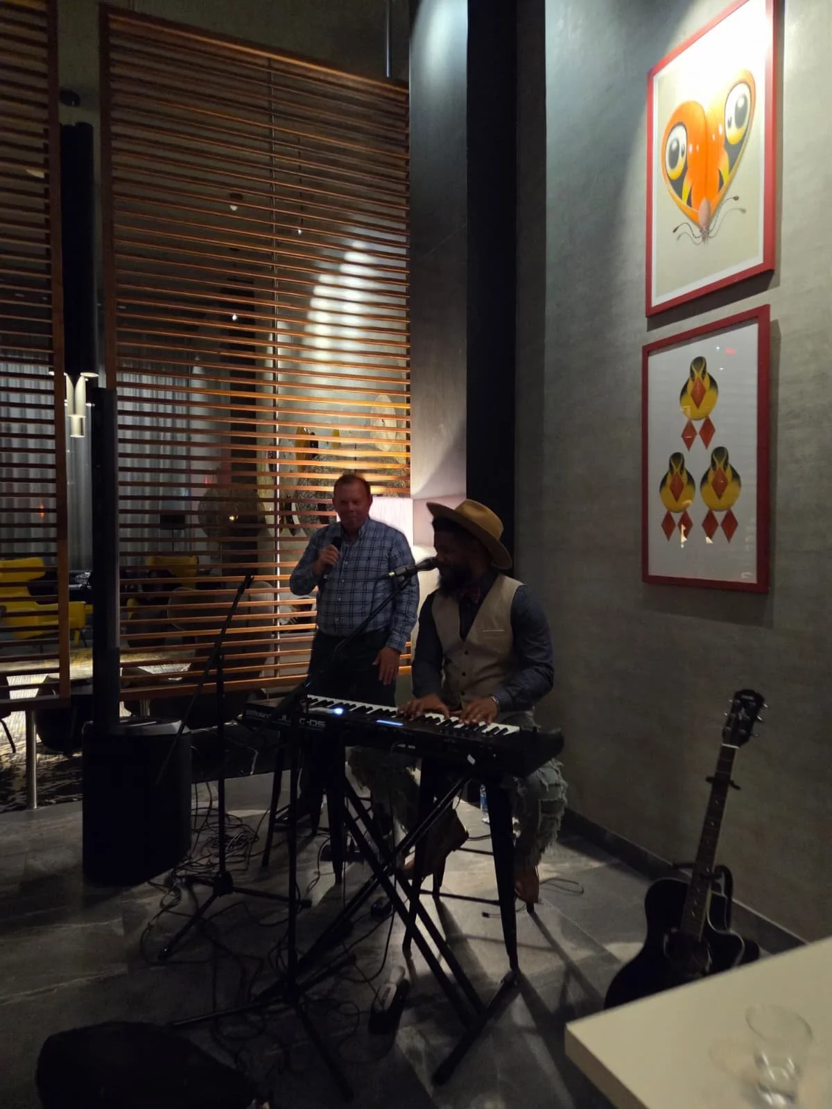 Musicians performing with a keyboard and microphone in a restaurant or cafe setting, with artwork on the wall and a guitar on the floor.