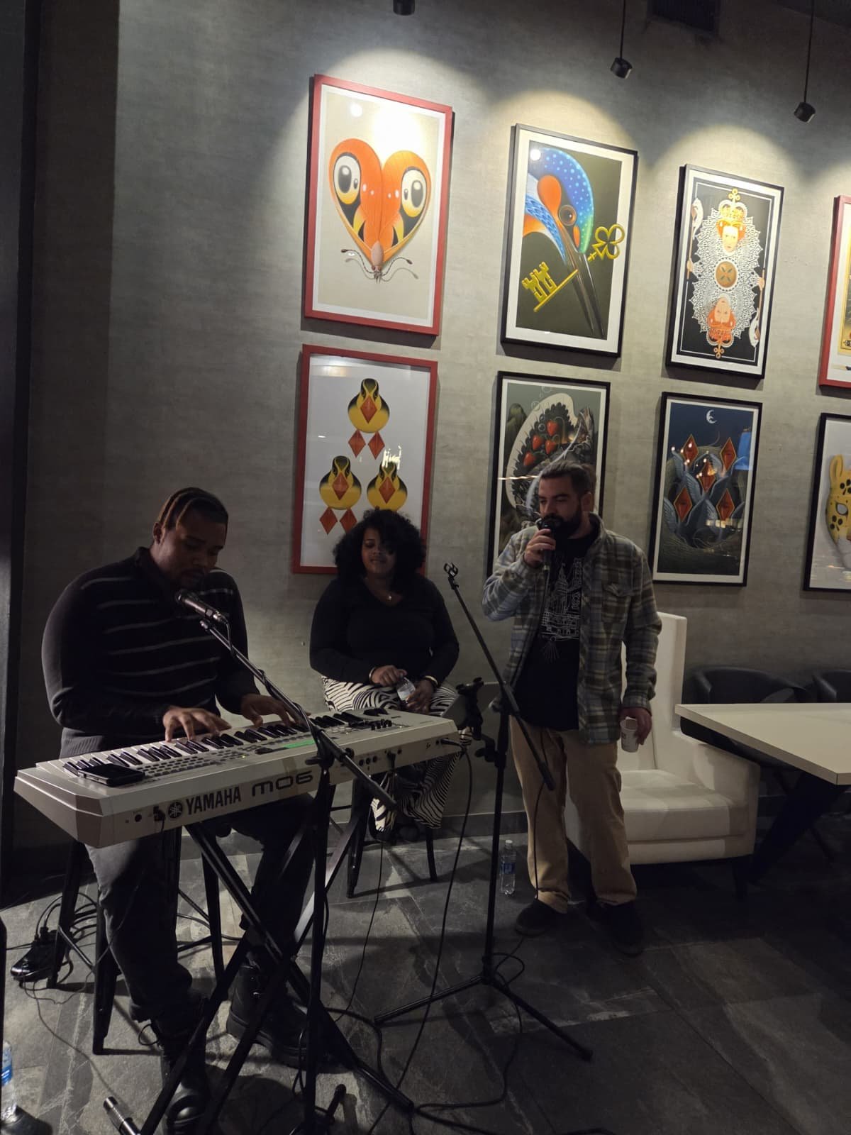 Three musicians performing in a room with art on the walls. One is playing a keyboard, one is singing into a microphone, and a woman is sitting and observing. The room has framed artwork and ambient lighting.