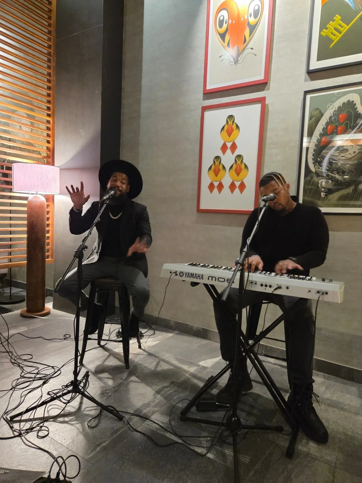 Two musicians performing indoors; one singing into a microphone and the other playing a Yamaha keyboard. They are seated in front of a wall decorated with colorful framed artwork.