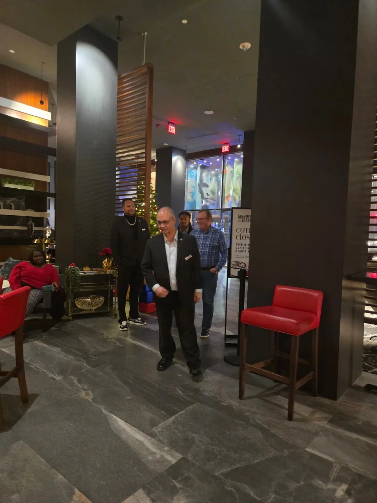 A group of four people, three men and one woman, are seen in a modern indoor setting, likely a restaurant or hotel lobby, decorated for Christmas with a small Christmas tree visible in the background. One man, dressed in a black suit, is walking forw