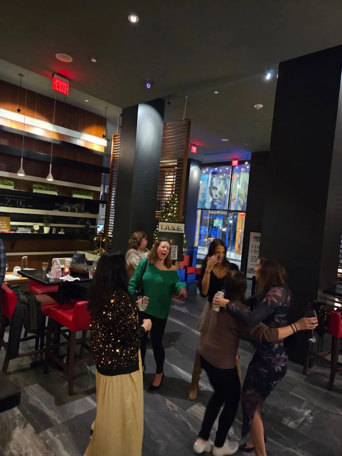 A group of women celebrating and laughing together in a restaurant or bar with modern decor, bar stools, and a Christmas tree in the background.
