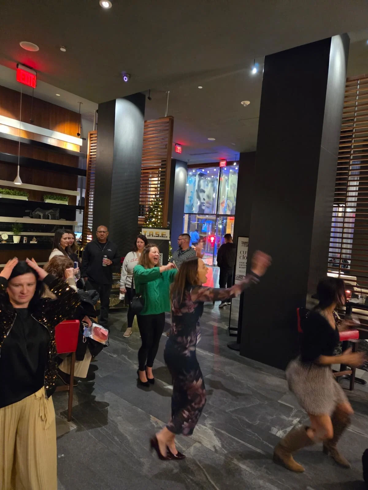 People dancing and socializing inside a modern, well-lit venue with dark walls, wooden accents, and large glass windows.
