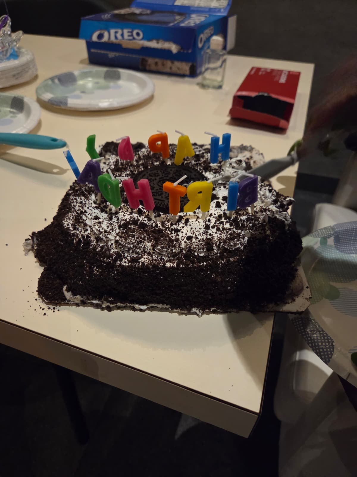 A chocolate birthday cake with white frosting and Oreo cookie crumbs, decorated with colorful birthday candles spelling out 'HAPPY BIRTHDAY' on top.