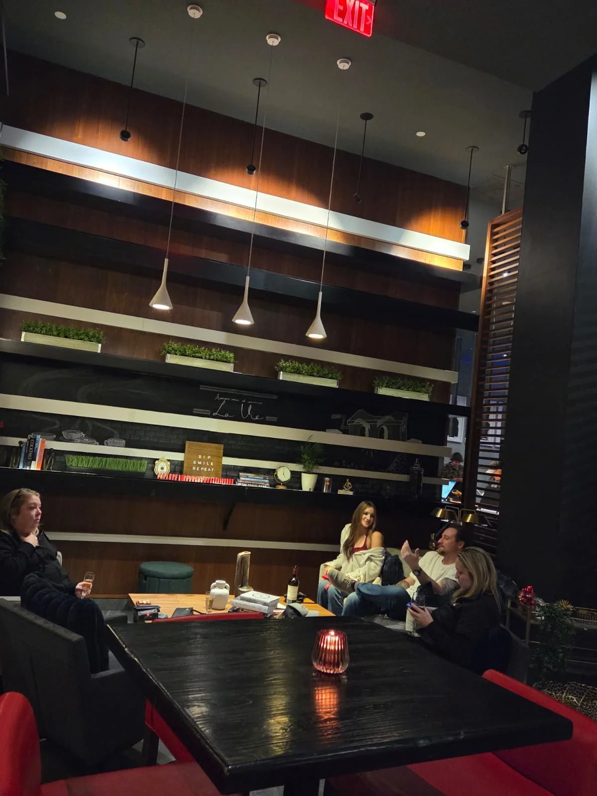 Group of four people sitting at a table in a modern indoor dining area with wood accents, pendant lights, a decorative shelf with books and plants, and a candle on the table.