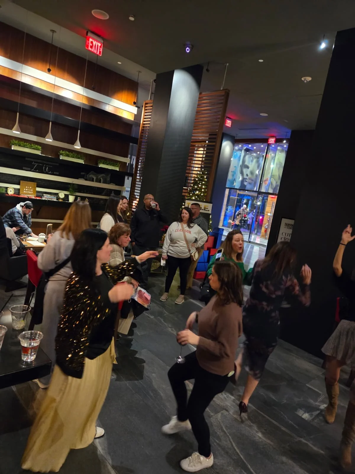 People dancing and socializing inside a modern restaurant or lounge. There is a bar area on the left and large glass windows in the background showing colorful lights and artwork. The atmosphere appears lively and festive.
