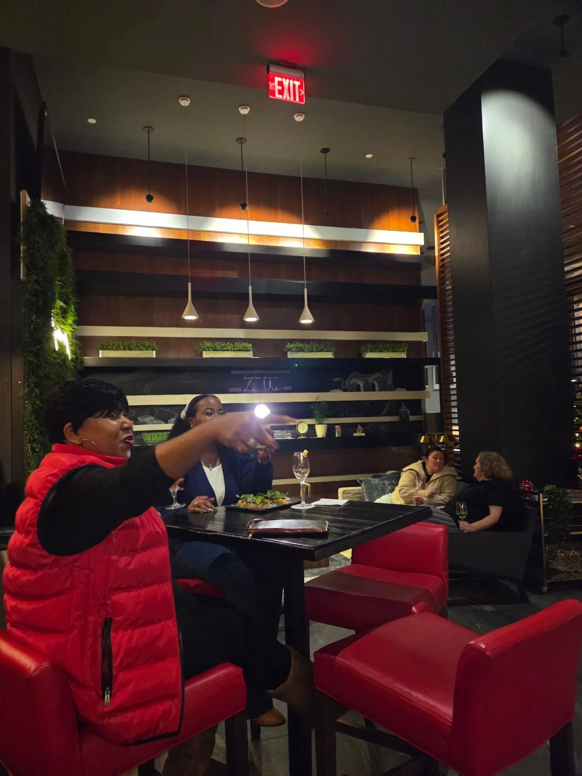 Inside a restaurant, a woman in a red vest is speaking and gesturing, with two women sitting at the table in the background. The decor includes wooden panels, shelves with plants, hanging lights, and a chalkboard wall.