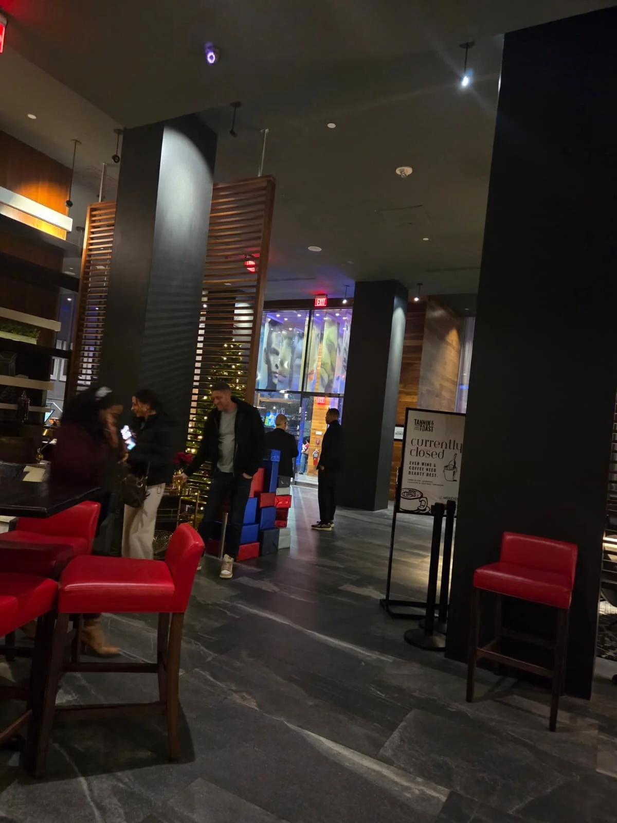 Interior of a restaurant or cafe with red chairs, dark floors, and modern decor. Several people are standing and talking, with a Christmas tree and large window visible in the background.