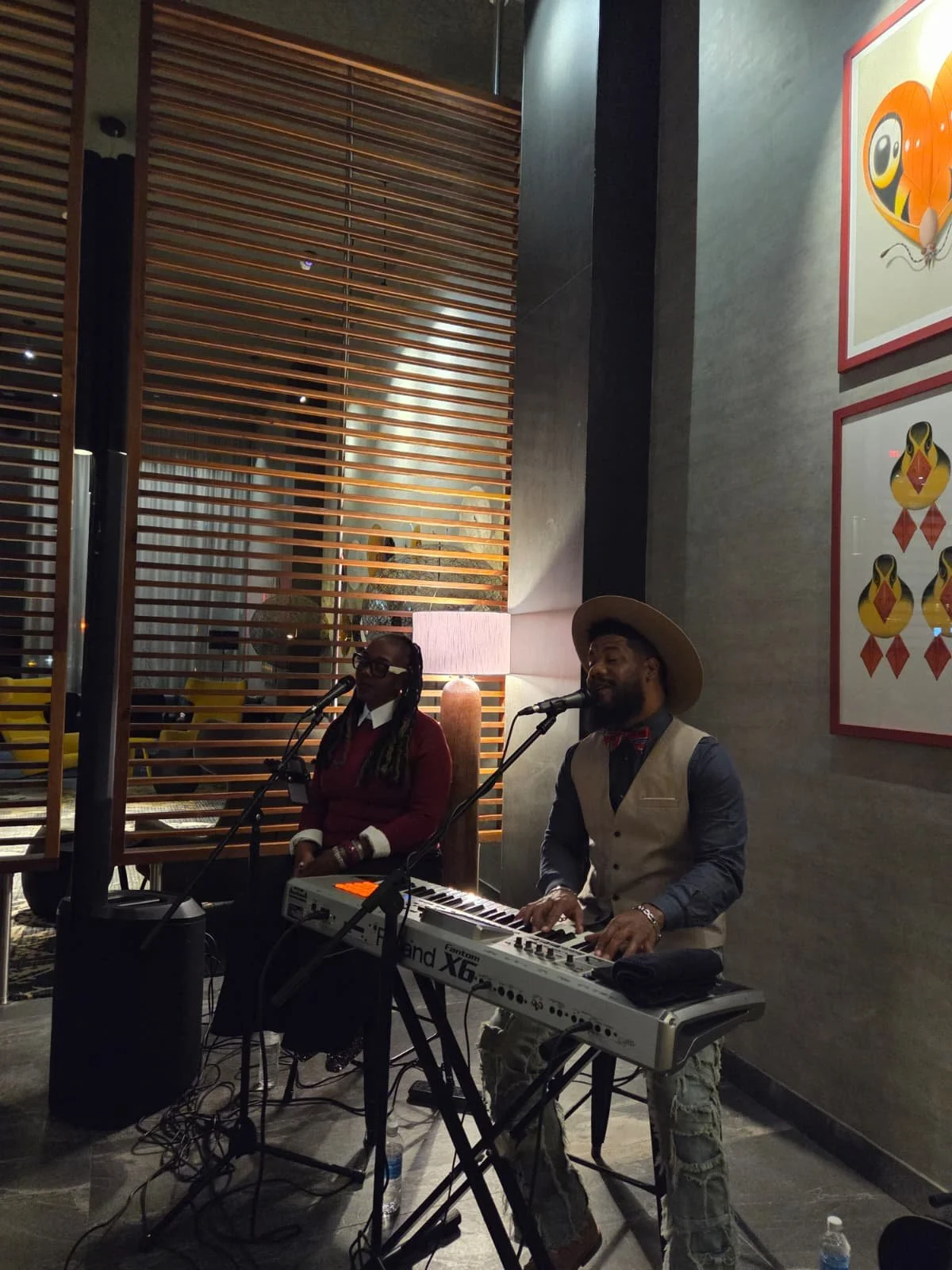 Two musicians perform with microphones indoors, one playing a keyboard and the other sitting with a microphone, in a modern venue with artwork on the wall.